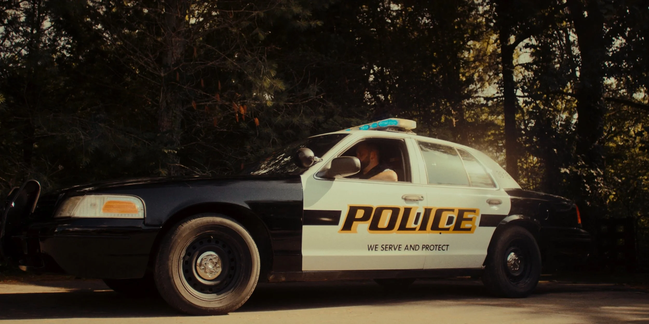 A black and white police car parked on a road, with a police officer inside. The vehicle is surrounded by trees and sunlight filtering through the branches.