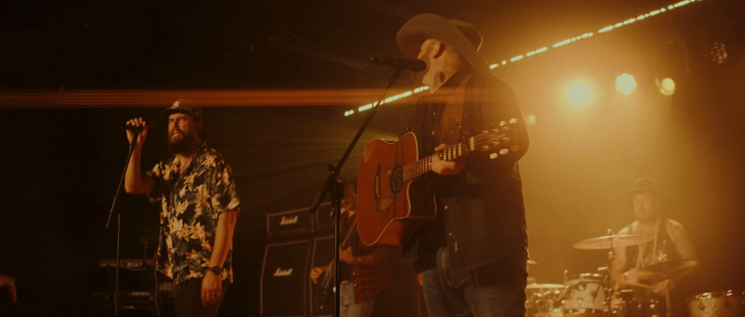 A band performing live on stage with three musicians visible: a singer with a beard wearing a cap and a floral shirt, a guitarist wearing a cowboy hat and sunglasses, and a drummer in the background. The stage is lit with warm yellow and orange light