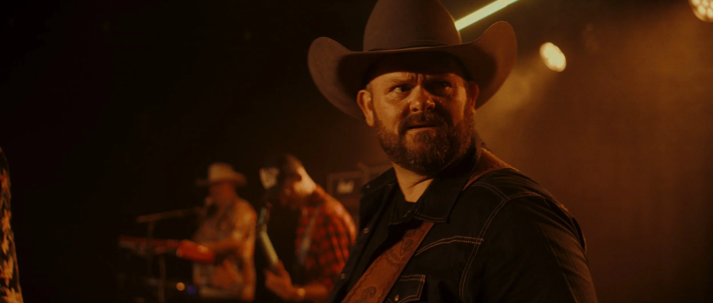 A man with a beard and mustache wearing a cowboy hat and black jacket looking over his shoulder in a dimly lit setting, with musicians in the background.
