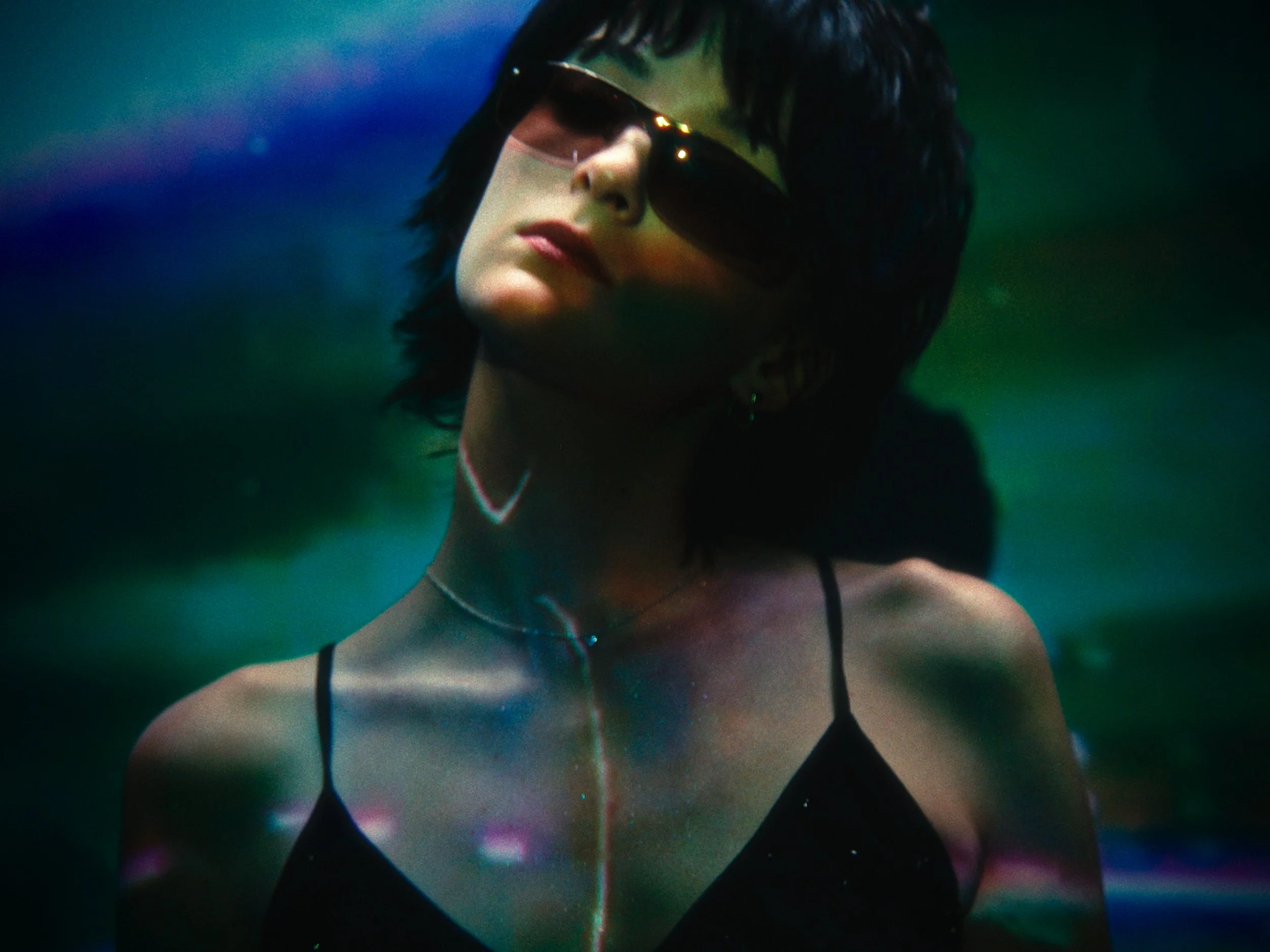 A woman with short dark hair and sunglasses in a dark, colorful setting.