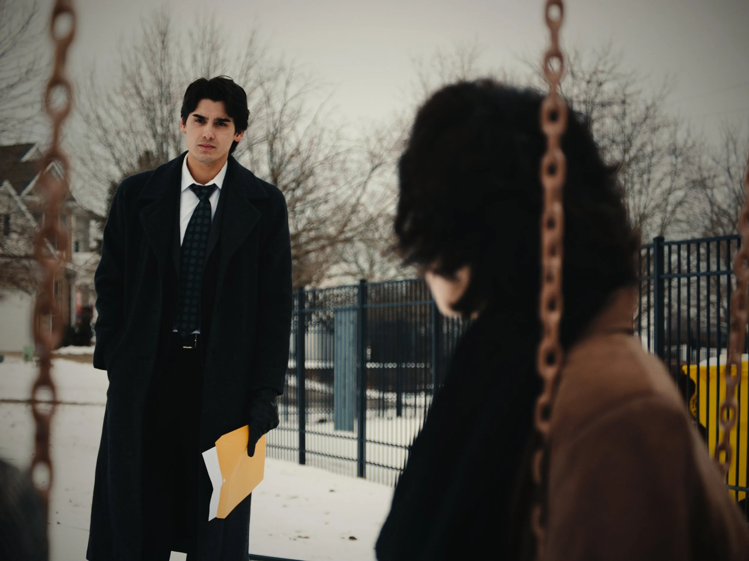 A young man with dark hair, wearing a black coat, white shirt, and navy polka dot tie, stands looking with a serious expression. He holds a yellow folder in his left hand. In the foreground, a woman with curly dark hair, wearing glasses and a brown c