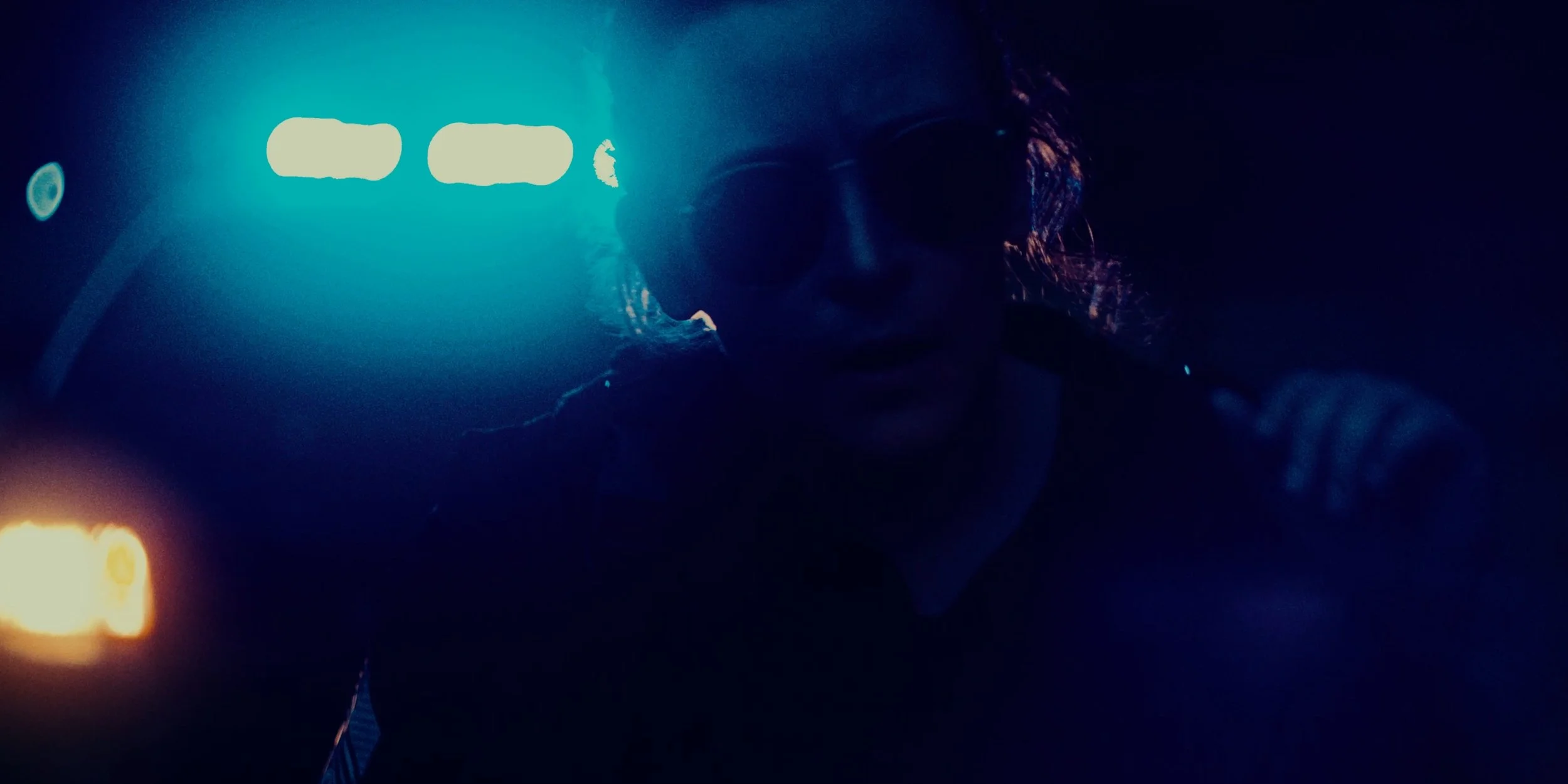 A person wearing sunglasses in a dark setting with neon lights and lens flares.