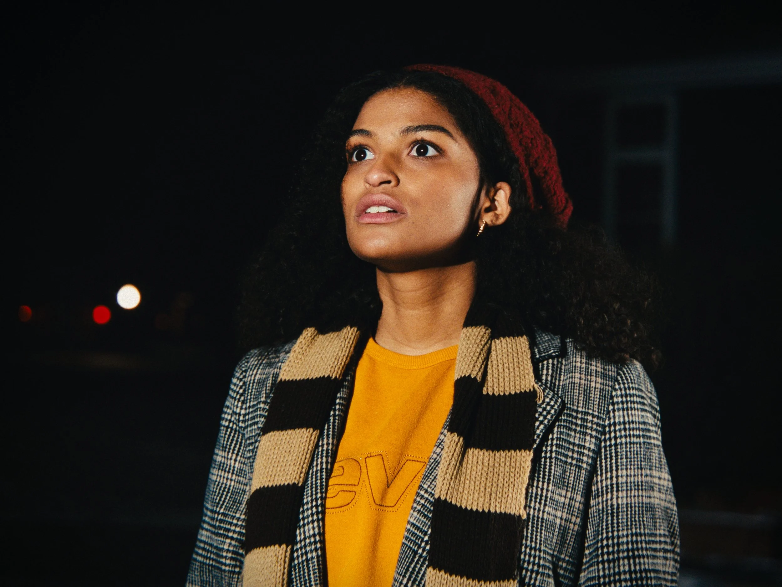 A woman with curly dark hair, wearing a red beanie, a checkered blazer, a yellow T-shirt, and a striped scarf, looks upward with a concerned or surprised expression against a dark background with some distant lights.