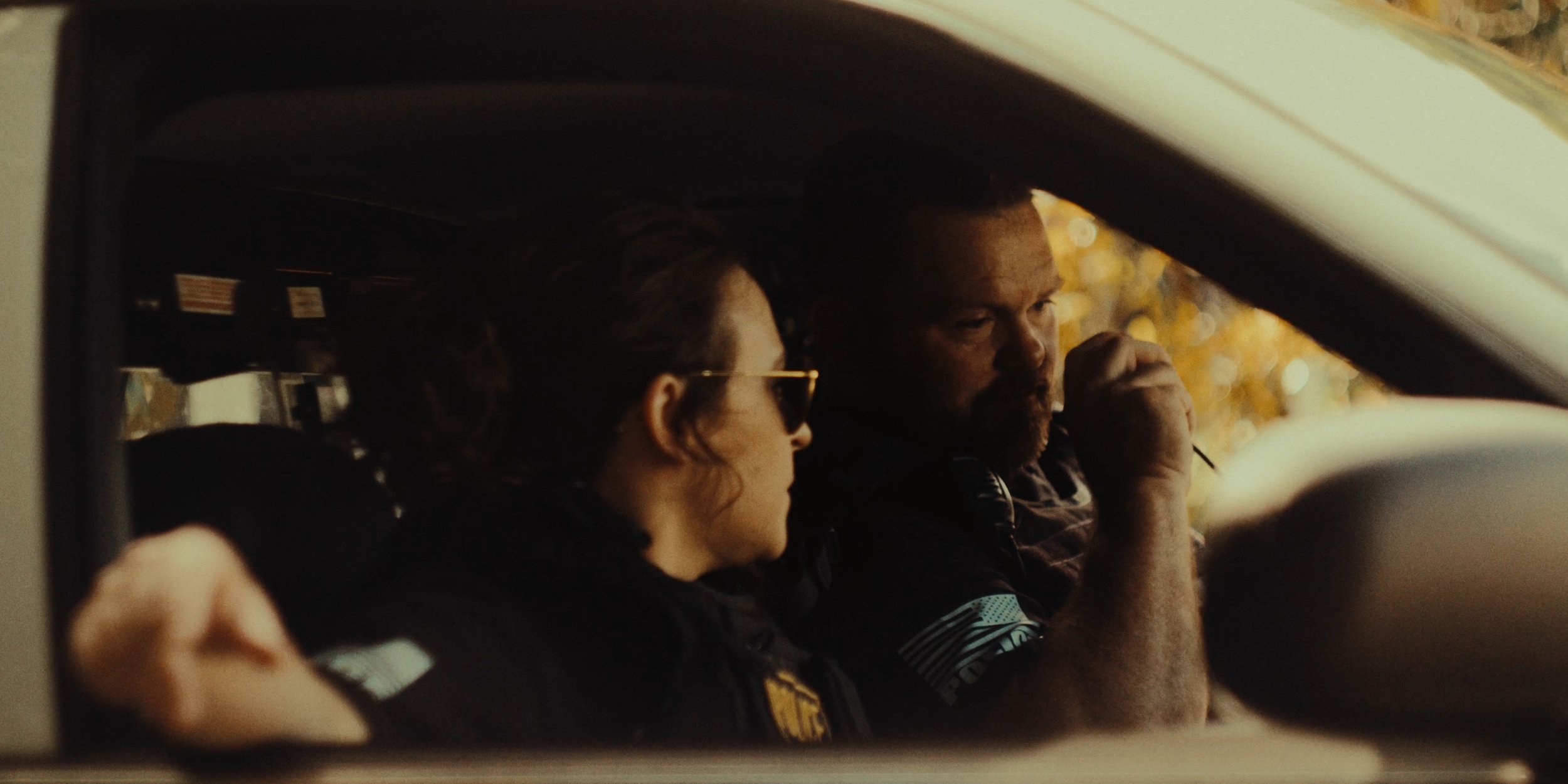 A woman with dark hair, sunglasses, and a black jacket and a man with a beard and short hair, appear to be talking inside a car, with the woman looking ahead and the man holding a cigarette near his mouth. The photo is taken through the car window.