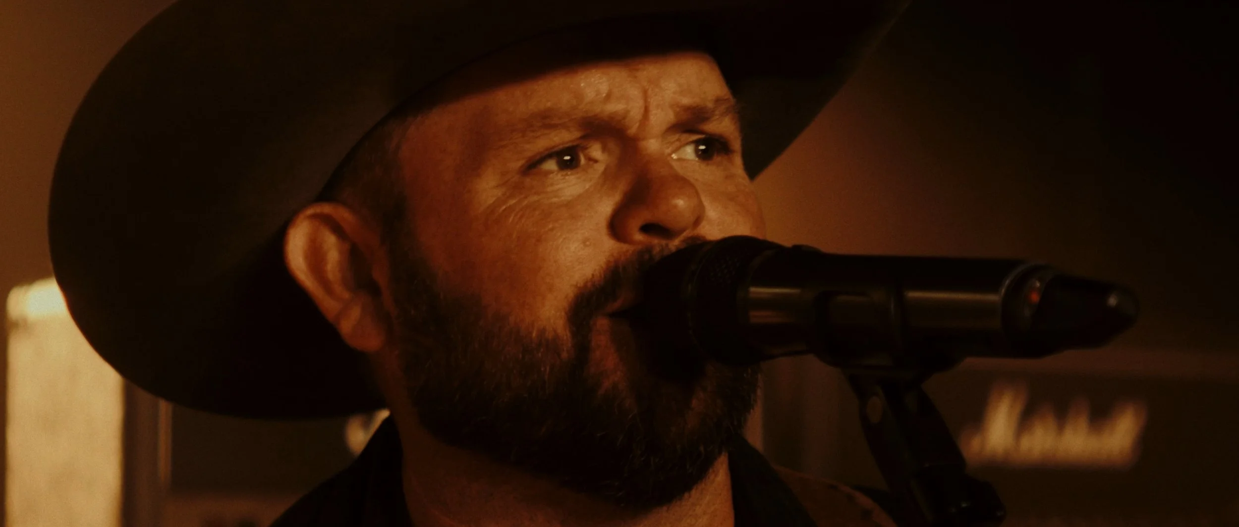 A man wearing a cowboy hat singing into a microphone in a dimly lit environment.