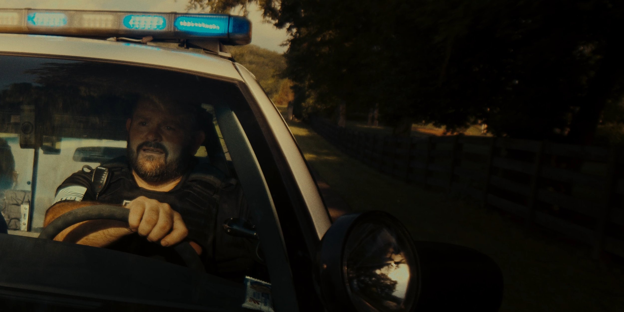 A police officer driving a patrol vehicle with a blue light bar on top during sunset, with trees and a fence visible in the background.