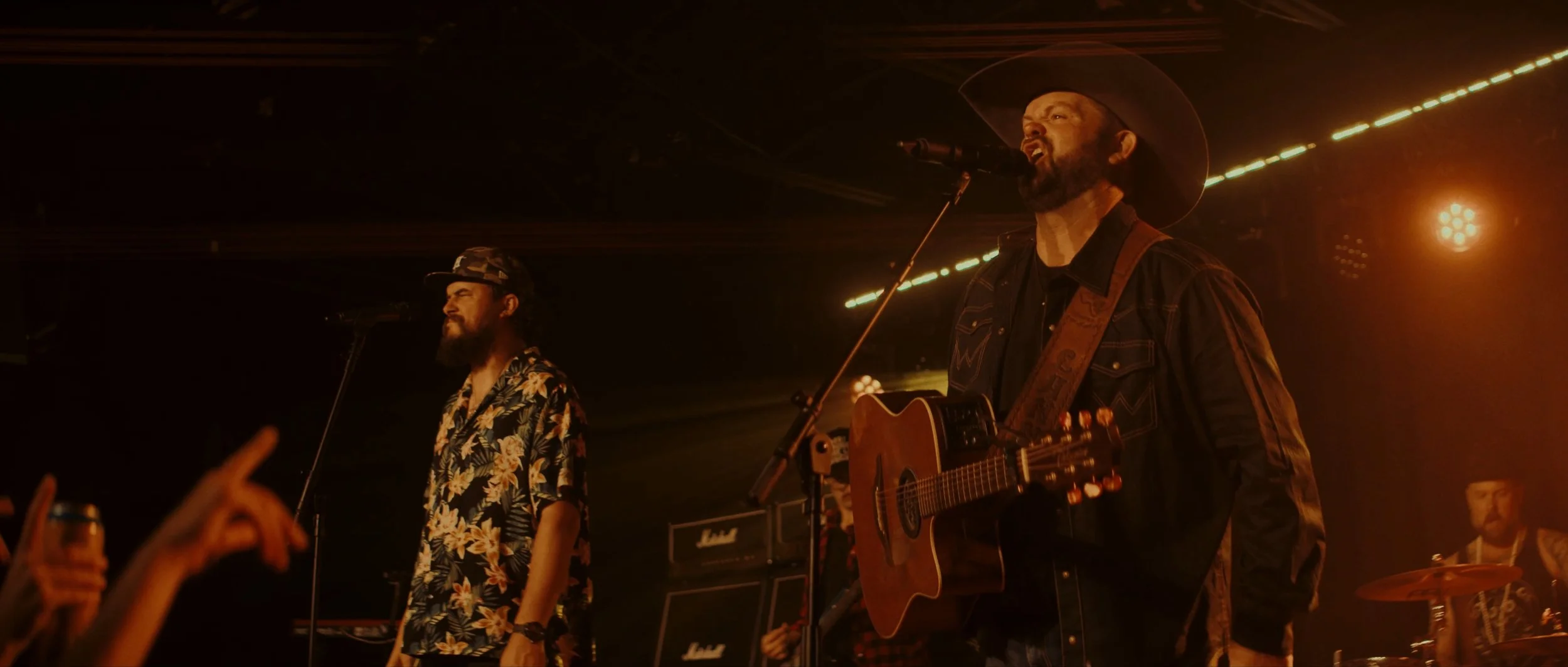 Music band performing on stage with warm lighting, featuring a vocalist with a beard and cowboy hat playing acoustic guitar, wearing a black shirt and denim vest, with other band members in the background, some in floral shirts, and audience raising 