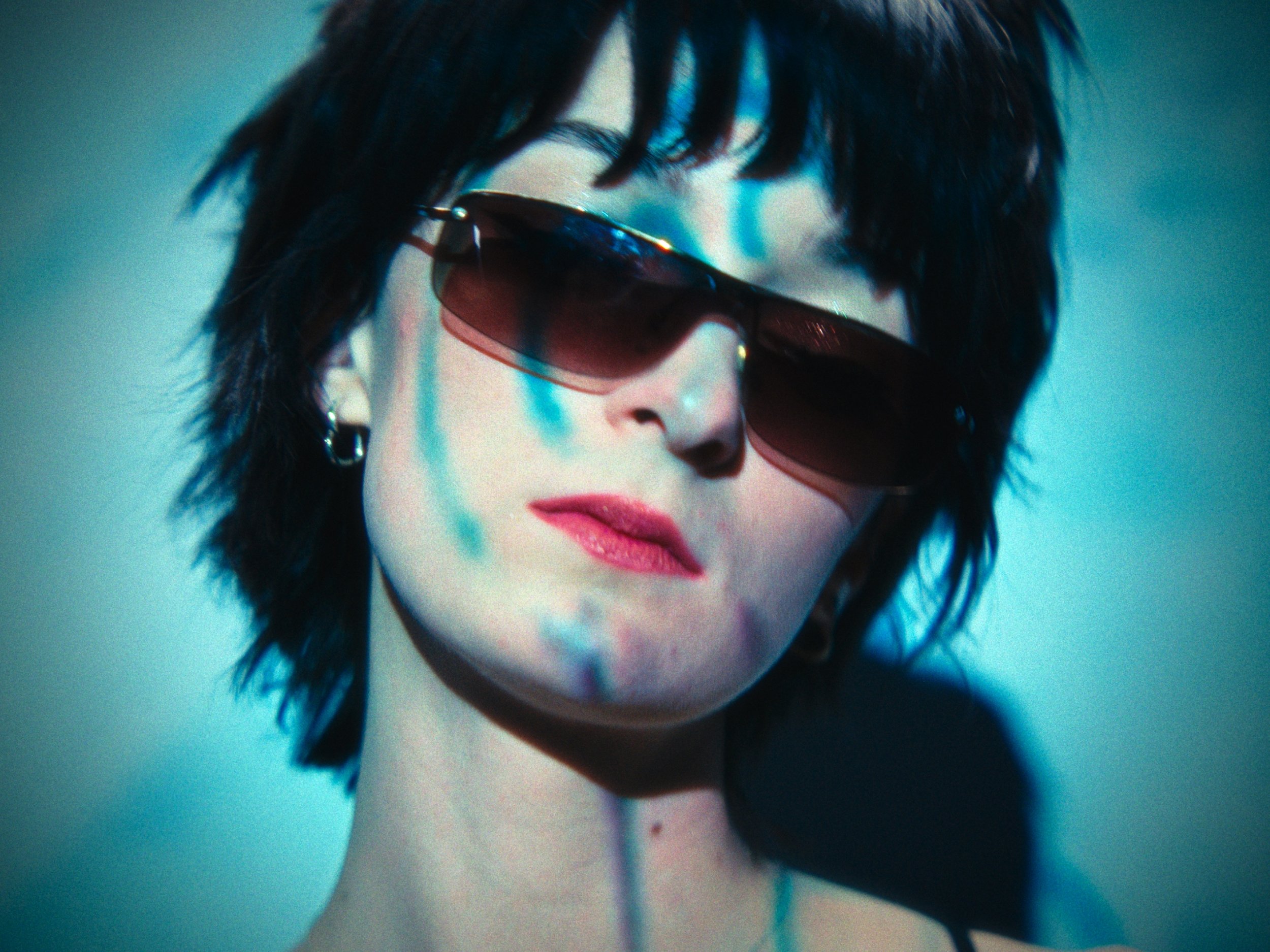 A woman with short black hair wearing sunglasses with a gradient tint, earrings, and red lipstick, illuminated with a mix of blue lighting.