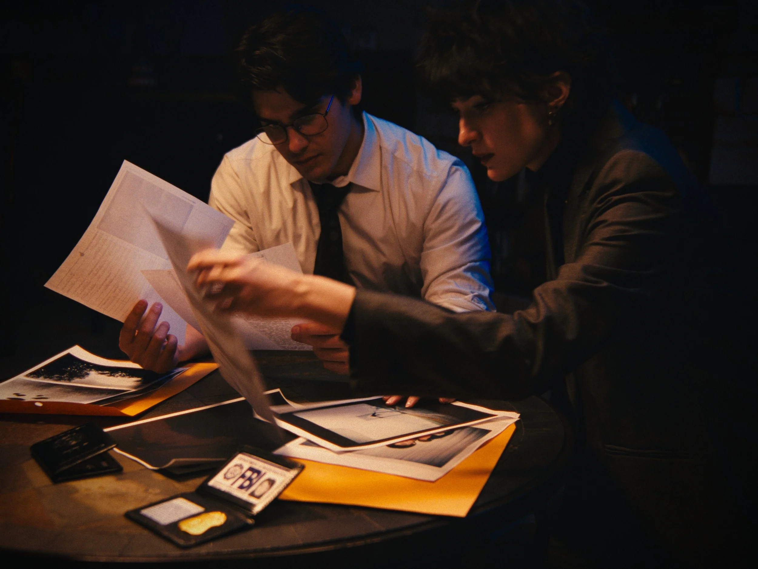 Two people, a man in glasses and a woman with short hair, are sitting at a table, examining documents. The table has photographs, a wallet, and other papers on it in a dimly lit setting.