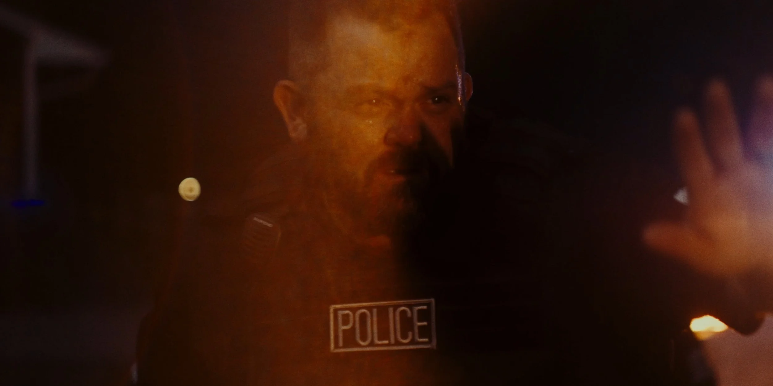 A police officer with a serious expression, wearing a uniform with a police badge, is inside a dark environment with a faint glow of lights.