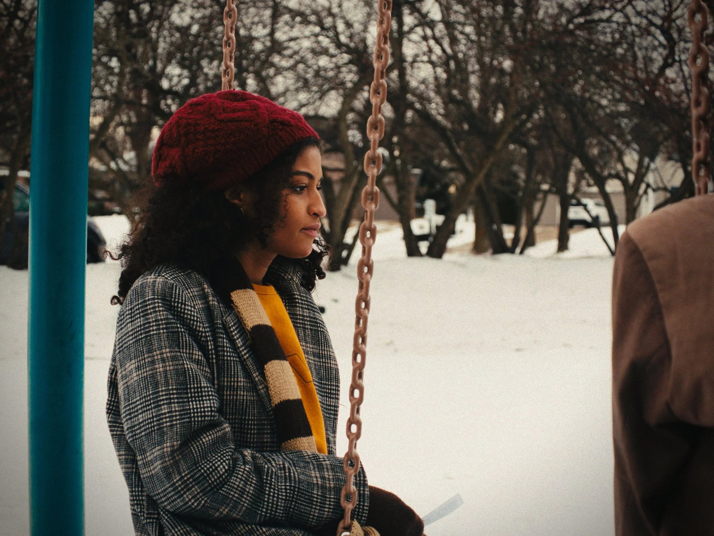 A woman with curly hair wearing a red knit hat, checkered blazer, and scarf sitting on a swing in a snow-covered park during winter.
