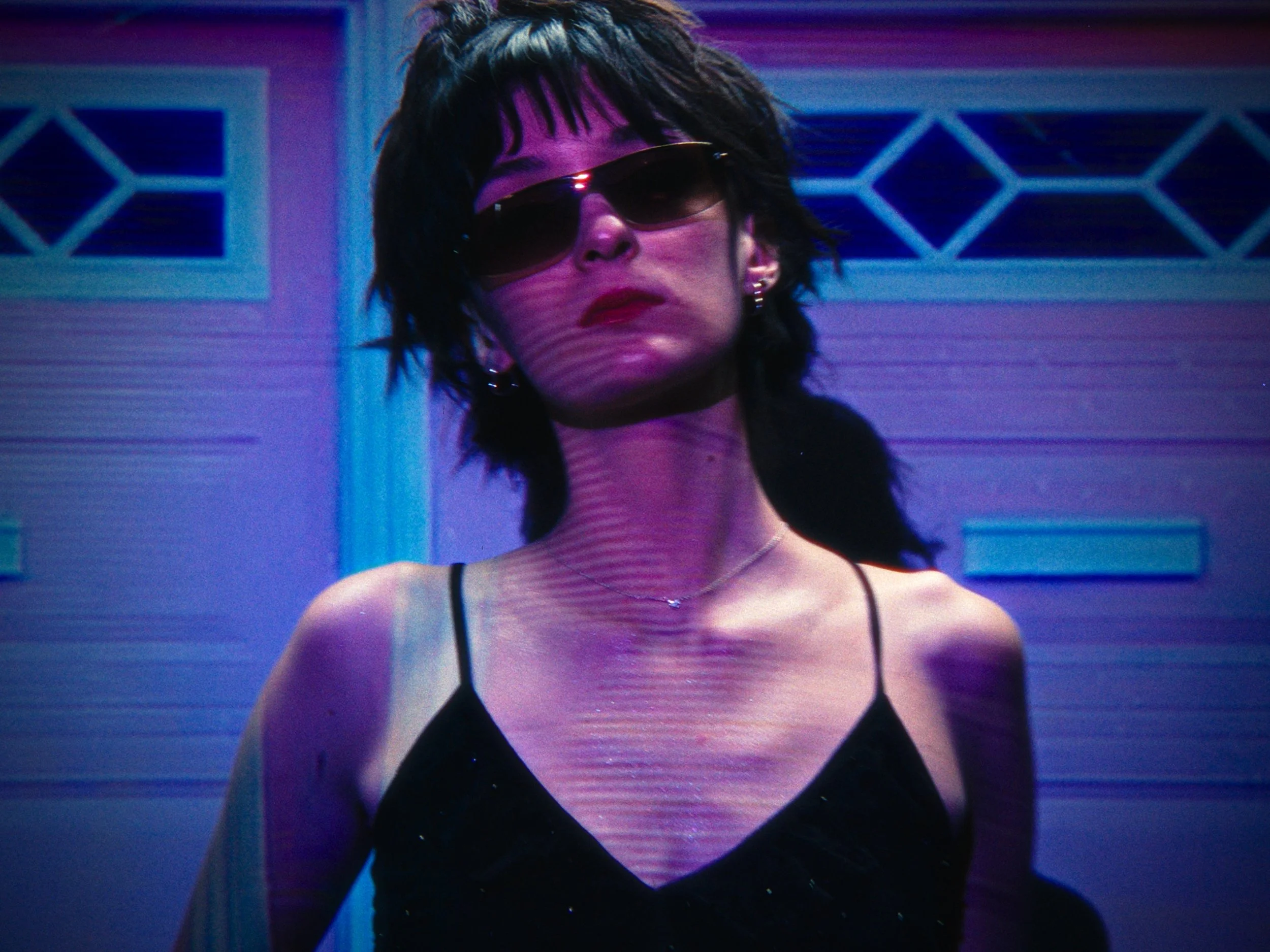 A woman with short black hair wearing sunglasses, a black spaghetti strap top, earrings, and a necklace, standing outdoors in front of a house with a garage door, illuminated by colorful purple and blue lighting.