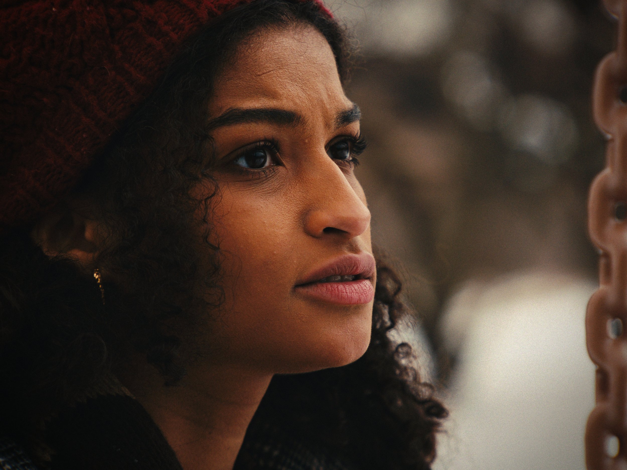Close-up of a young woman with curly hair, wearing a red knit hat, looking thoughtfully to the side.