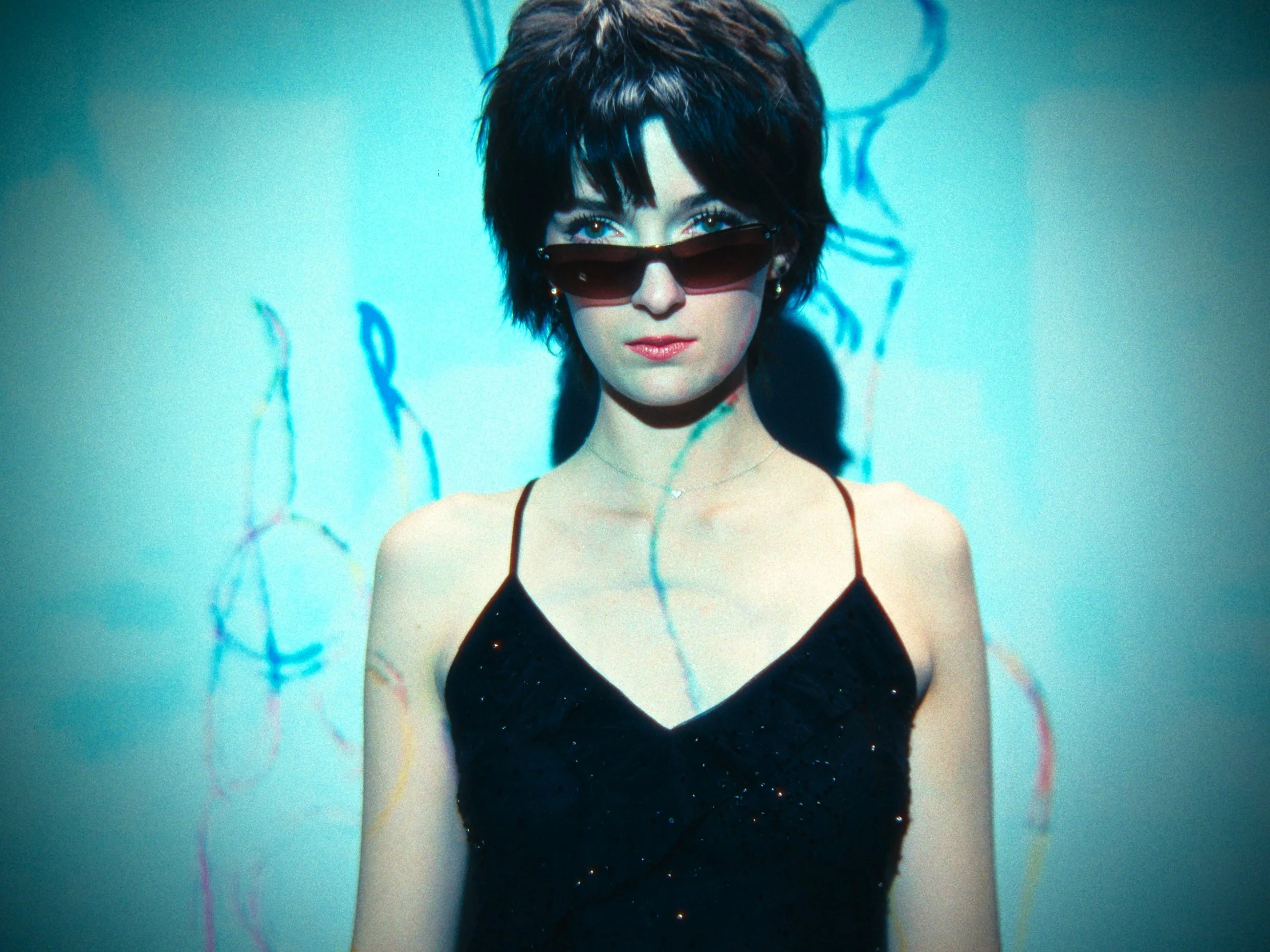 A woman with short dark hair and sunglasses posing against a turquoise background with colorful abstract lines.