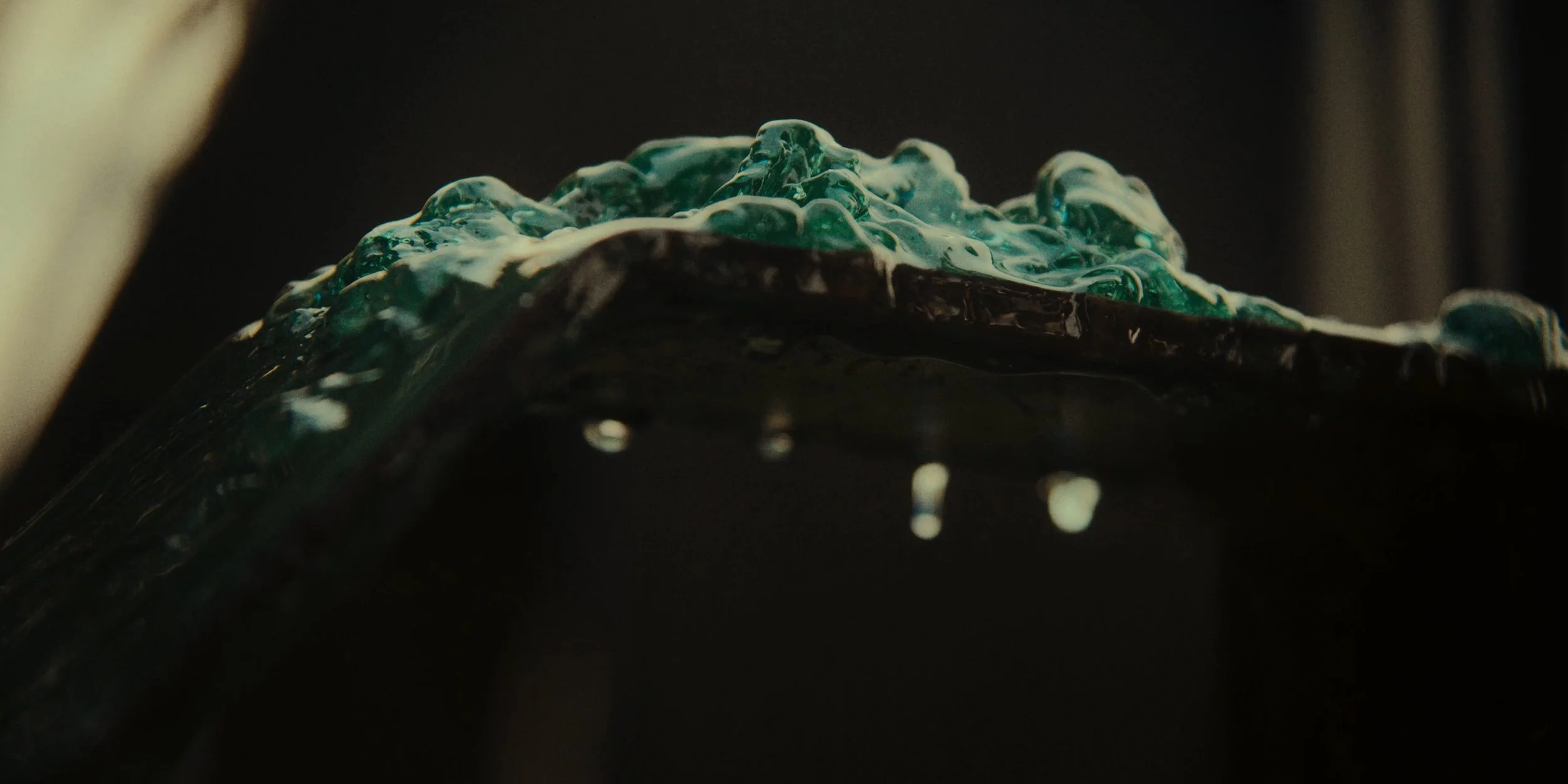 Close-up of a dark, flowing, textured liquid with a greenish tint over a black surface, with some small droplets beneath.