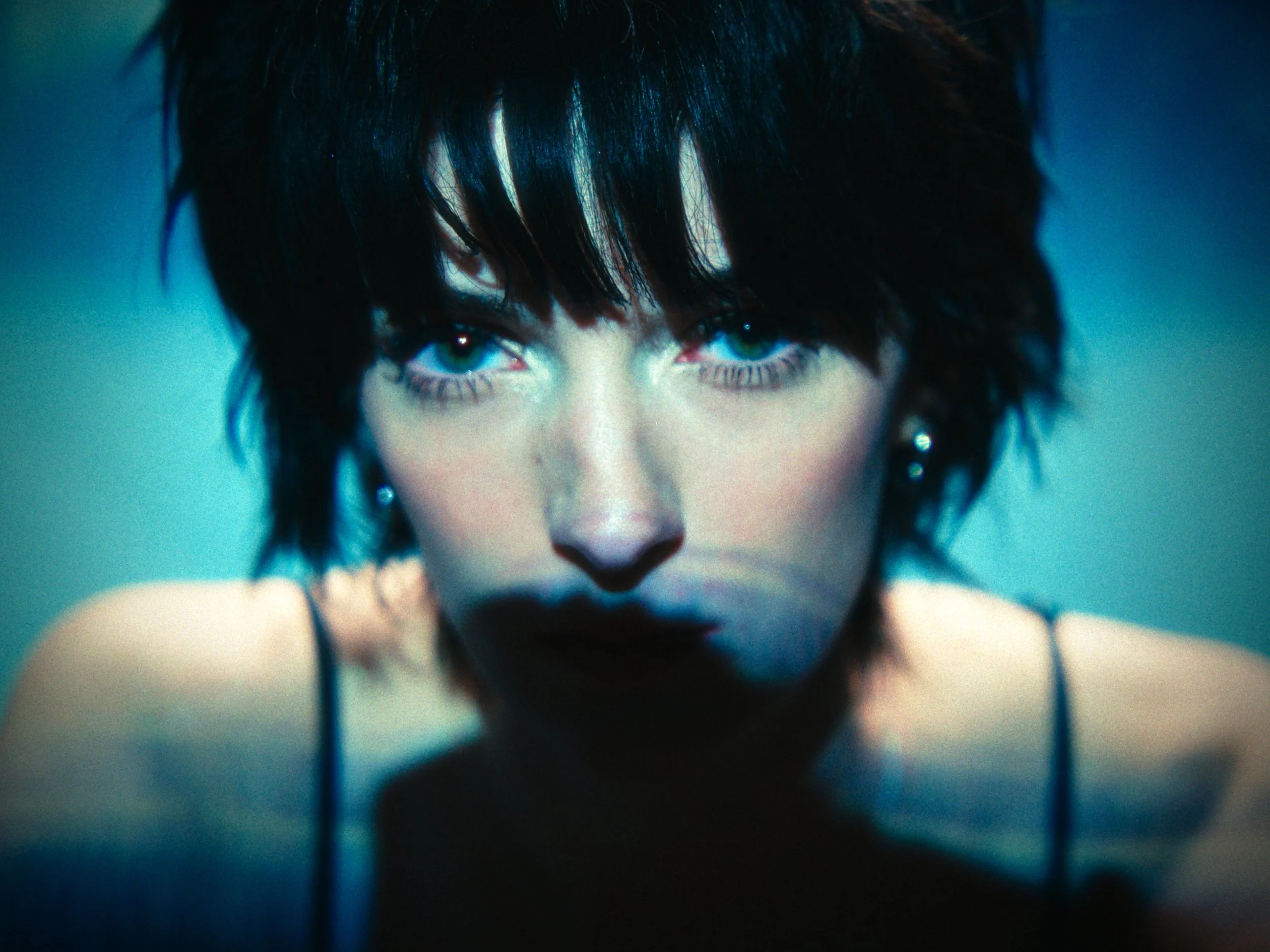 Close-up of a woman with dark, short, tousled hair, intense blue eyes, and dark makeup, wearing a thin strap top.
