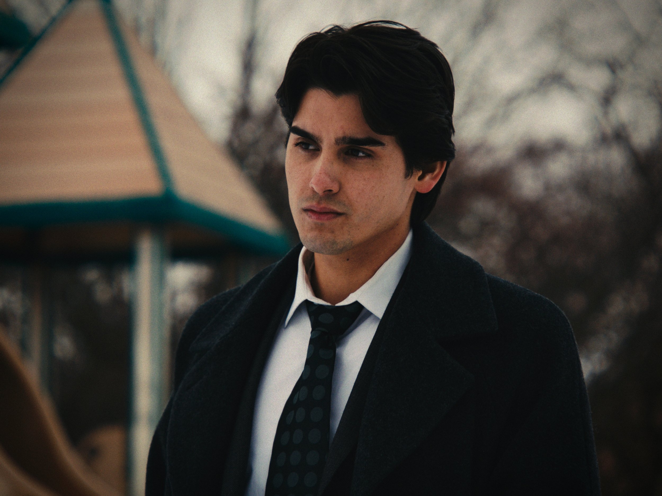 A young man with dark hair, wearing a white shirt, black coat, and a black bow tie with polka dots, standing outdoors near a playground on a cloudy day.