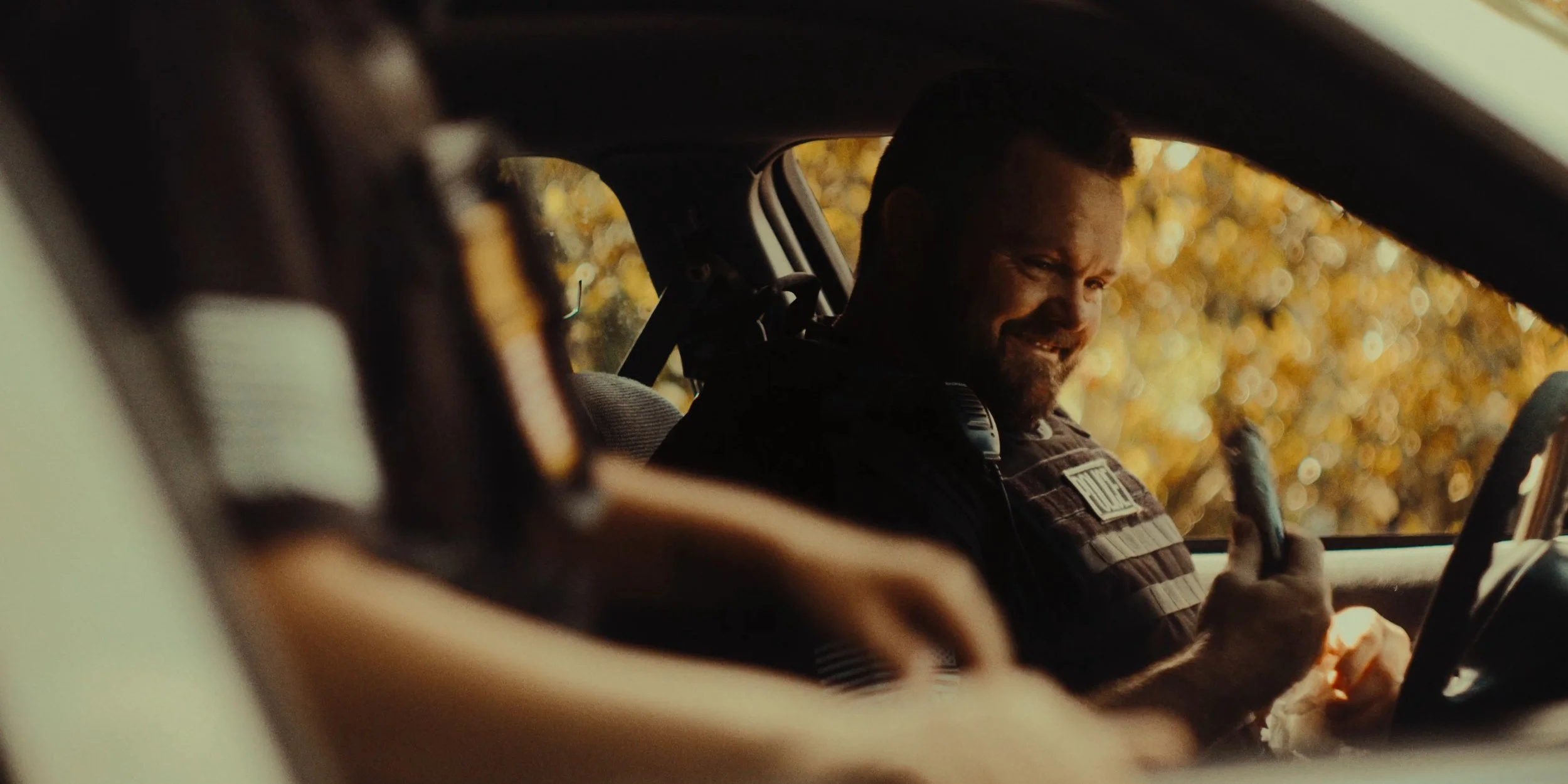 A smiling police officer inside a patrol car, looking at a handheld device, with autumn-colored trees visible outside the window.