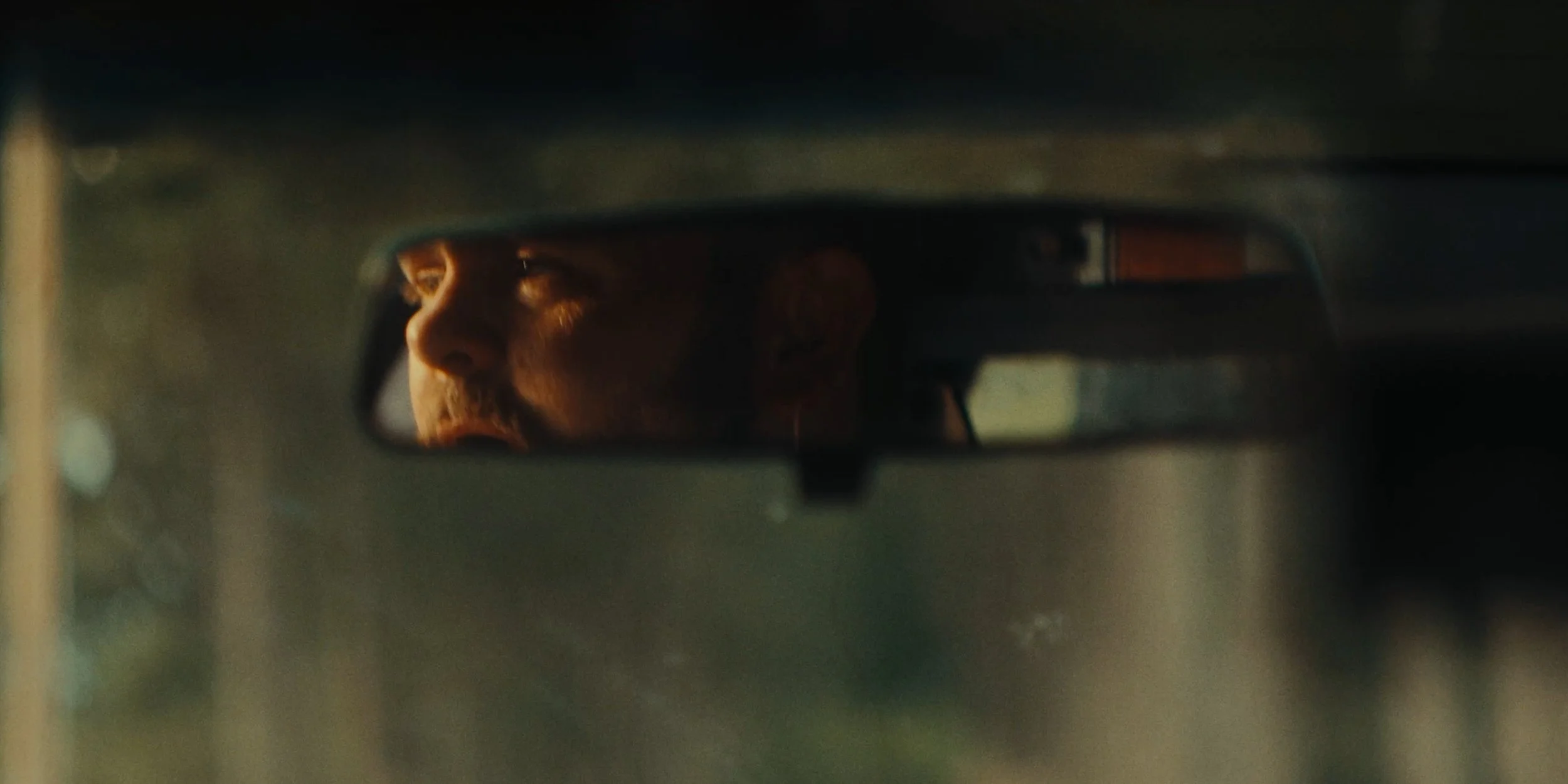 Close-up of a man’s face reflected in a car’s rearview mirror, appearing contemplative and partially illuminated.