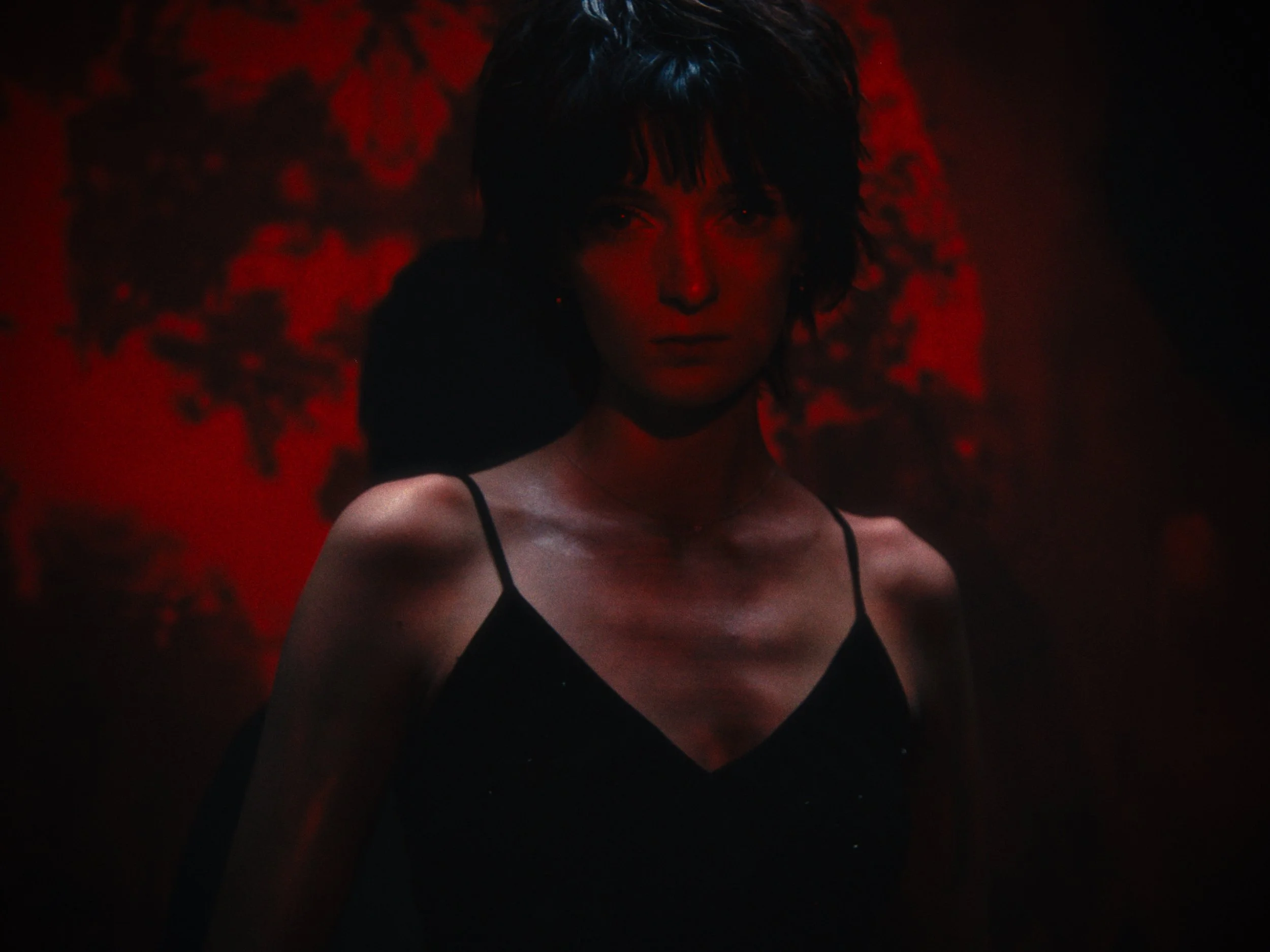 Woman with short dark hair in a black spaghetti strap top, illuminated by red lighting, with dark backgrounds.