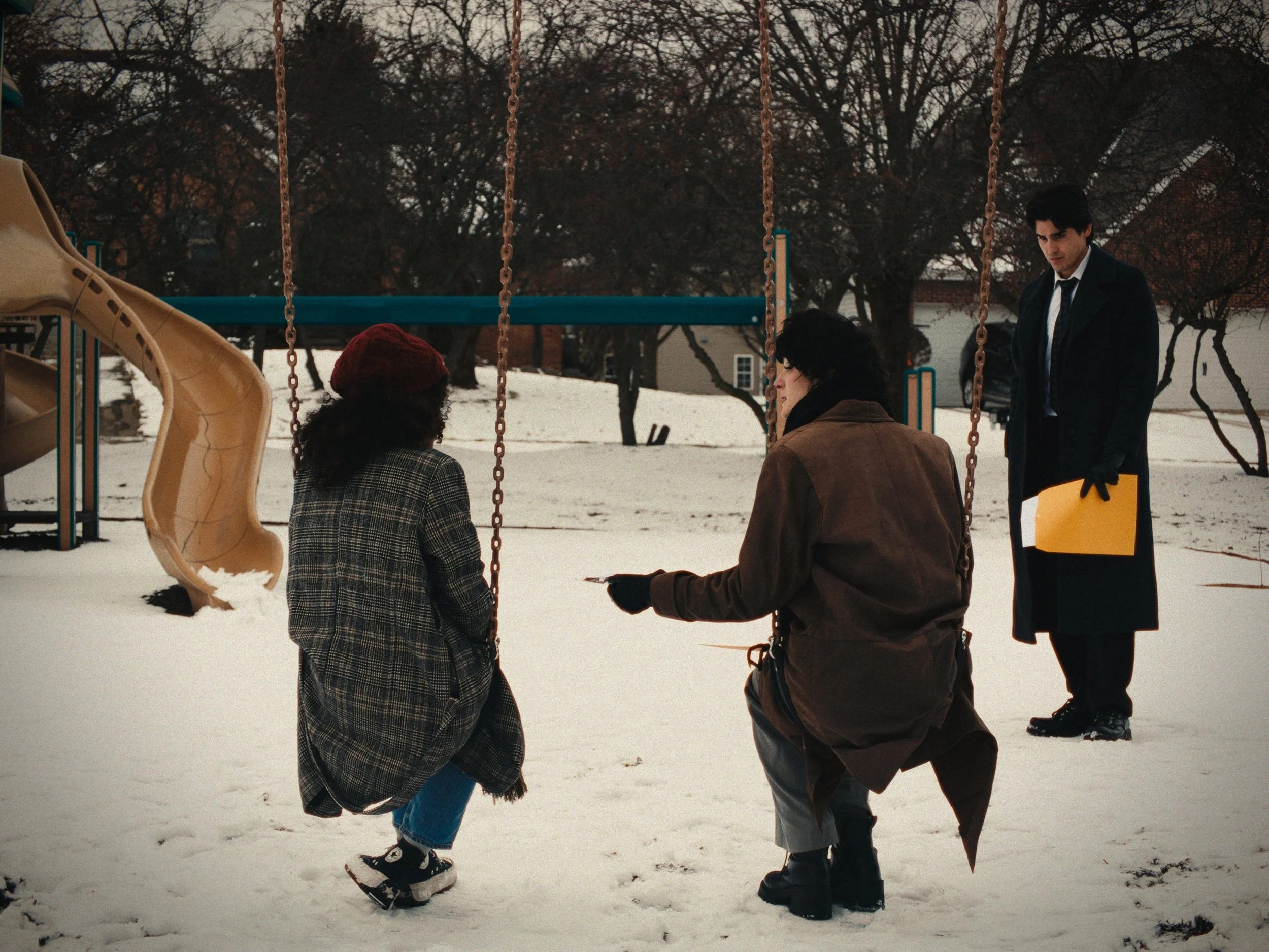 Three people in a snow-covered playground, two squatting and one standing, with a slide and trees in the background.