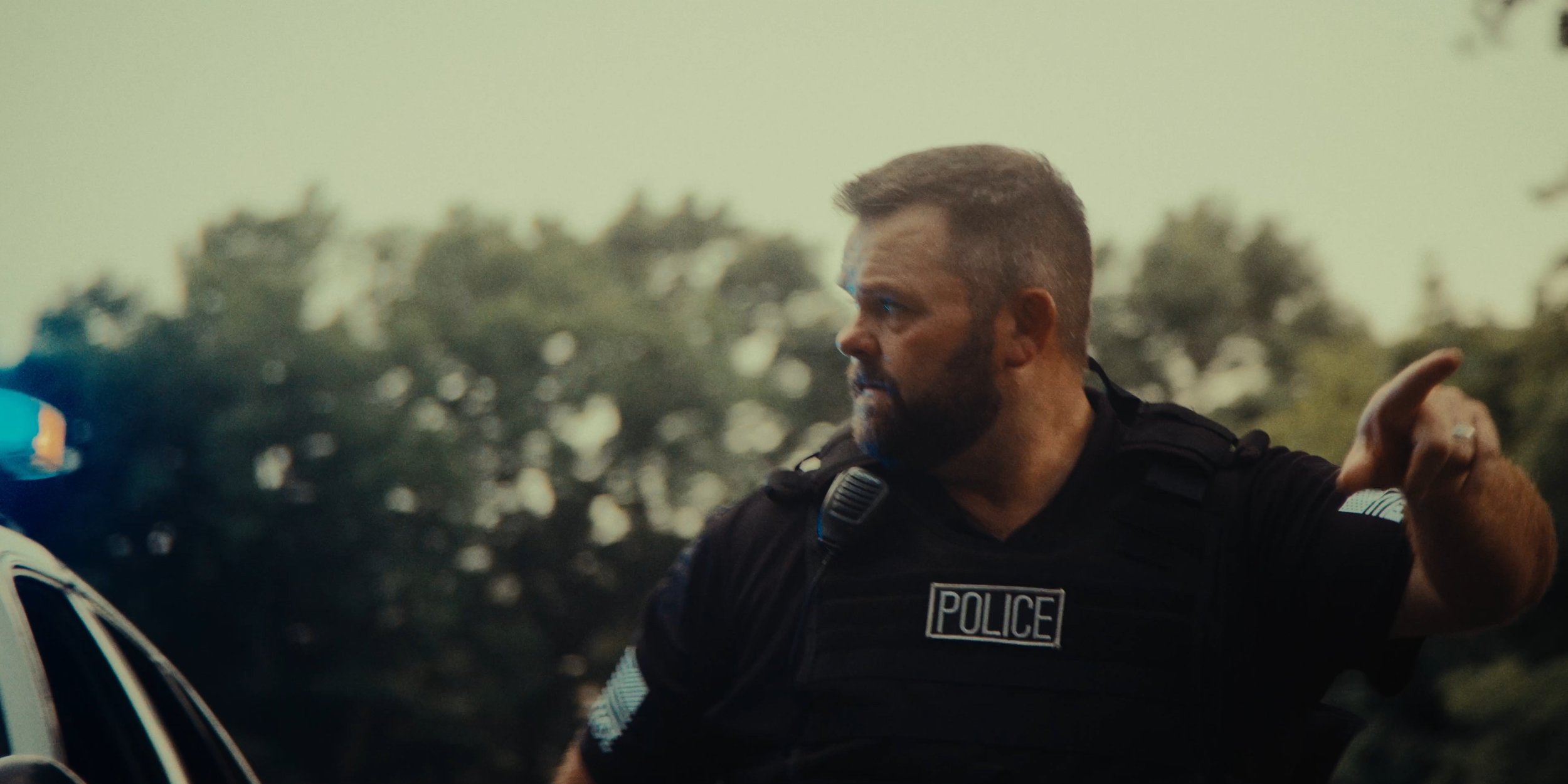 A police officer with a beard, wearing a black uniform with a police badge, is pointing with his right hand, with trees in the background.