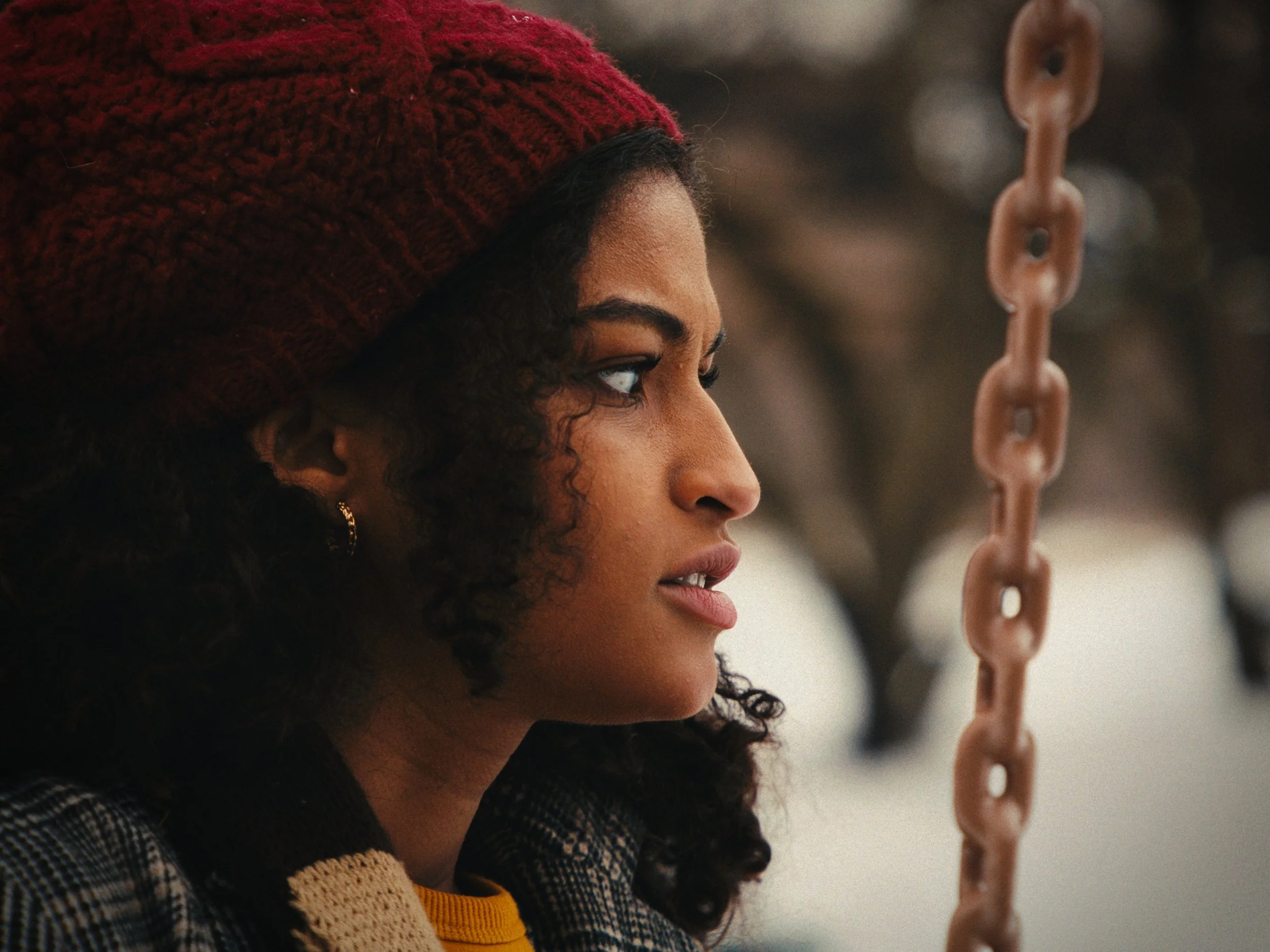 Side profile of a young woman with curly hair, wearing a red knit hat and earrings, looking at a chain hanging in front of her.