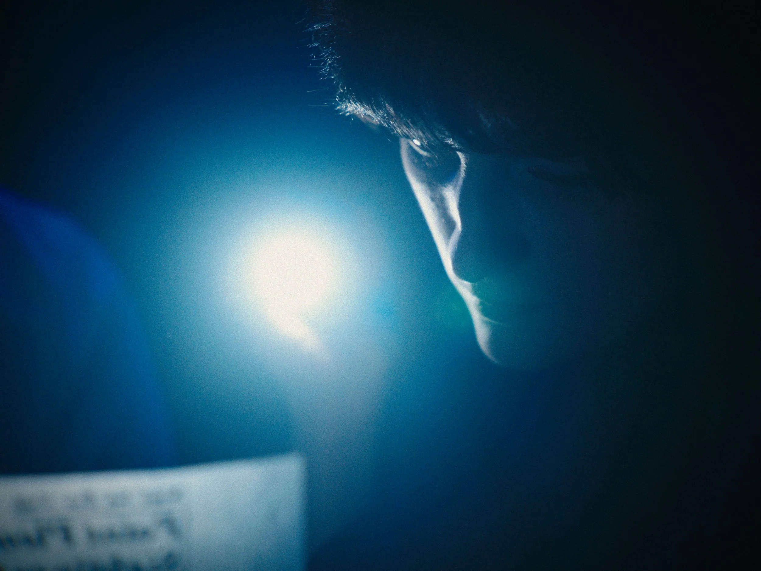 Close-up of a person's face, partially lit by a bright blue light, with a serious expression and dark hair.