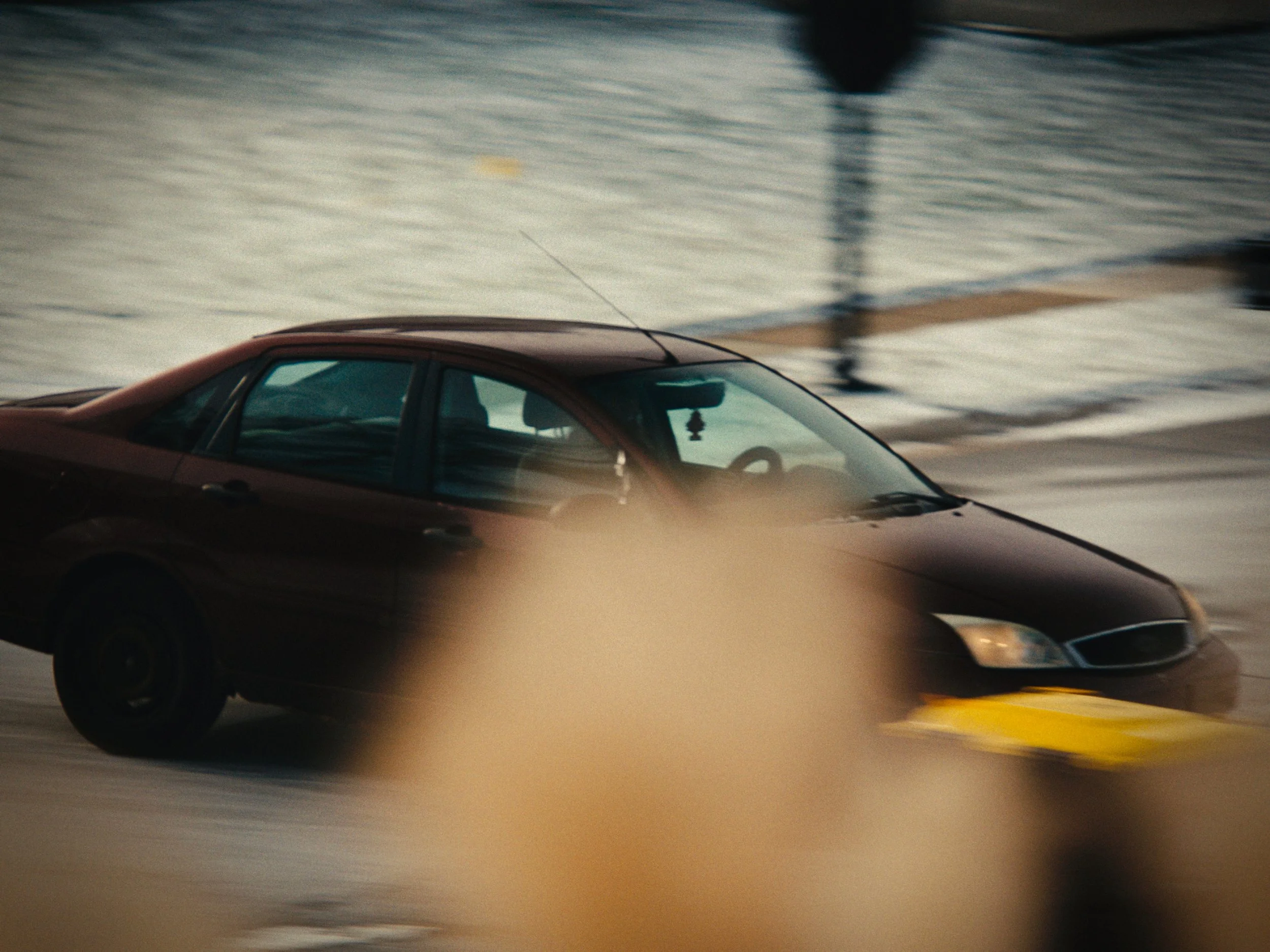 A dark-colored sedan driving on a wet street with visible reflections, blurred in motion, during daytime.