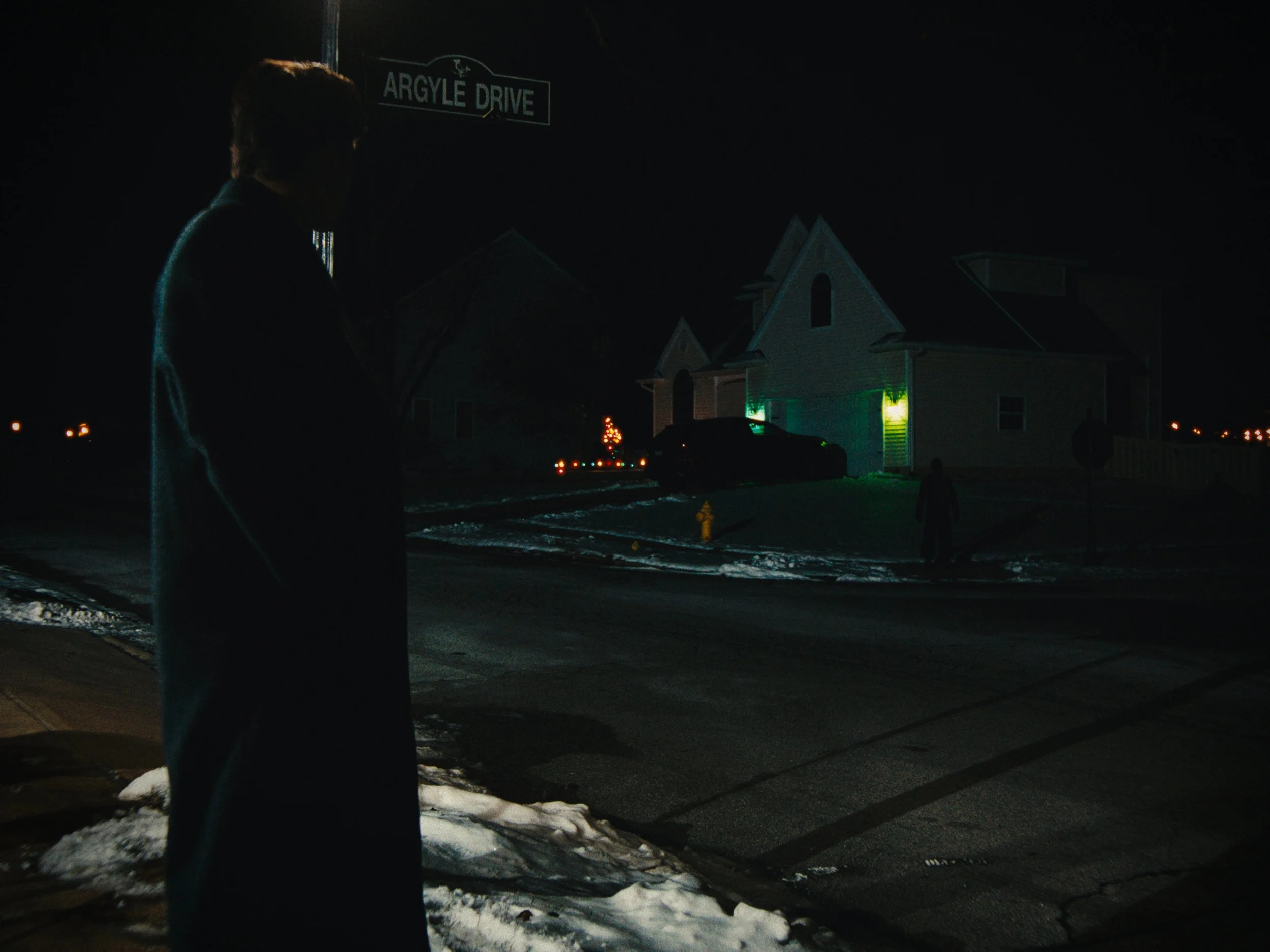 Nighttime scene at the corner of Argyle Drive with a person standing on the sidewalk, looking towards a house illuminated with green and yellow lights and a Christmas tree in the yard.
