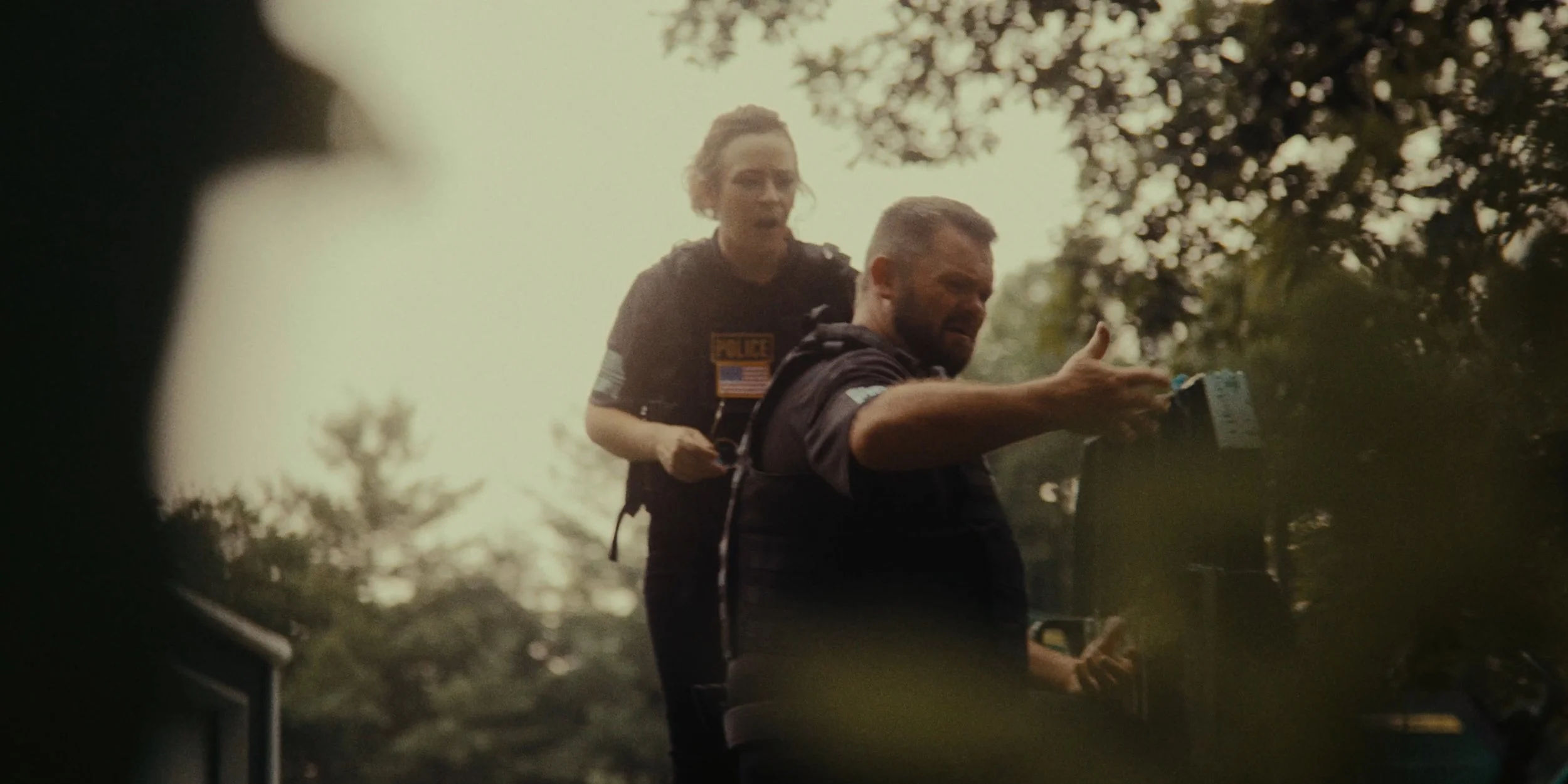 A man with a beard and a woman with curly hair, both in police uniforms, outdoors in a wooded area during daylight. The woman is sitting on the man's back.