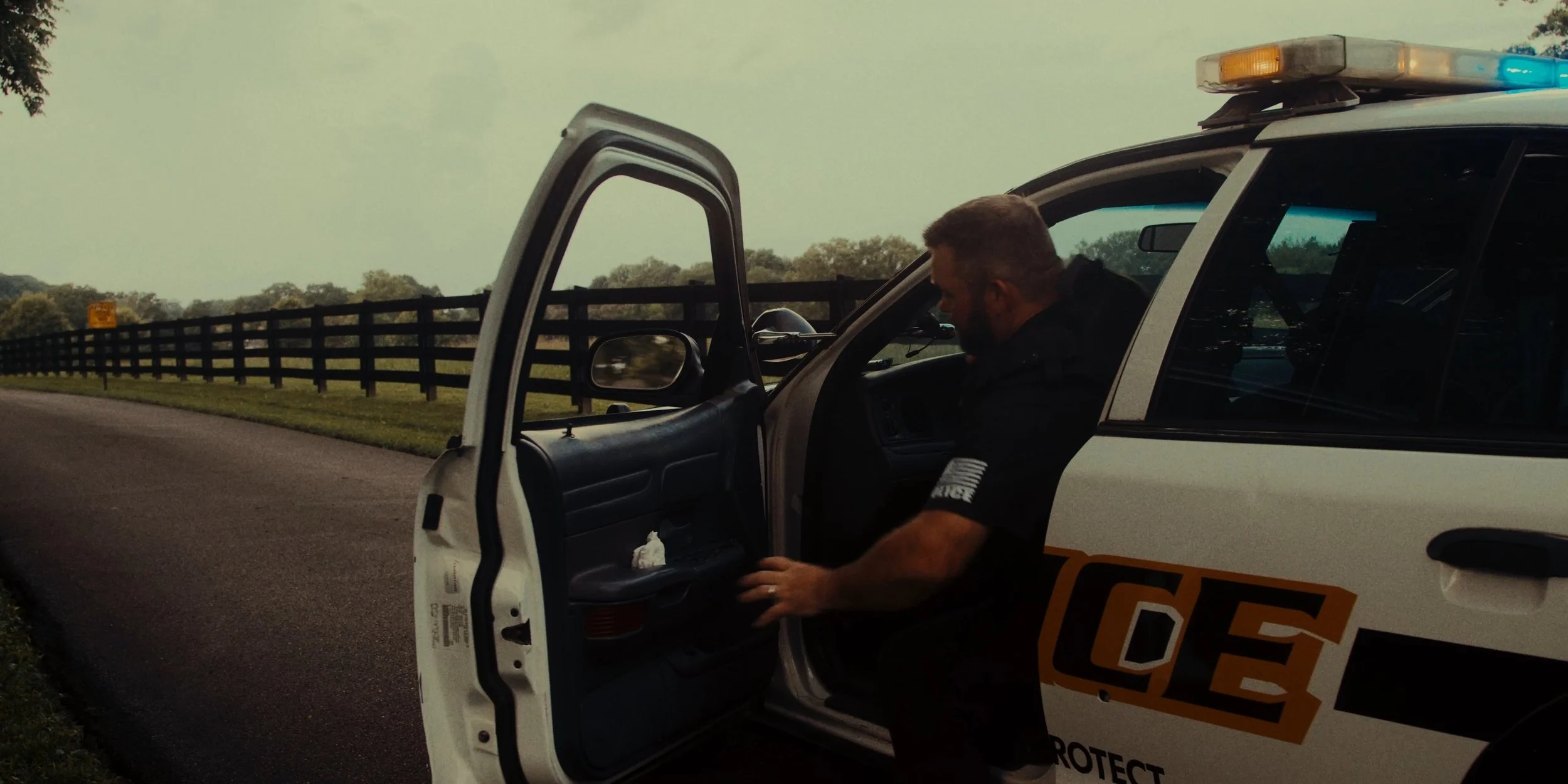 A police officer with short hair and a beard focusing on equipment inside a police vehicle, parked along a rural road with a wooden fence and green fields in the background.