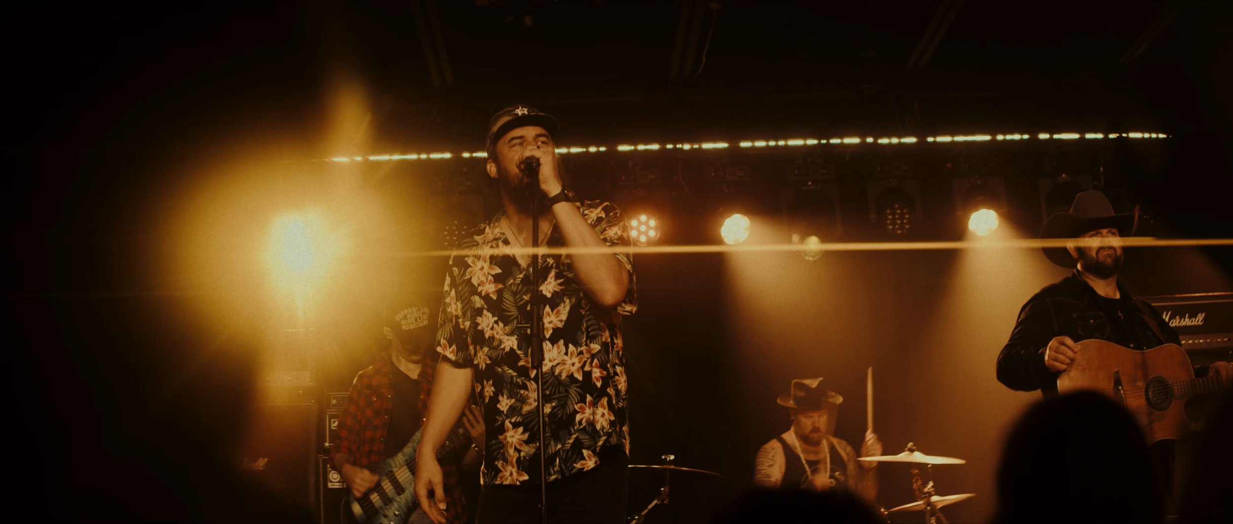Musicians performing on stage with warm lighting, including a singer in a floral shirt, a guitarist with a cowboy hat, and a drummer.