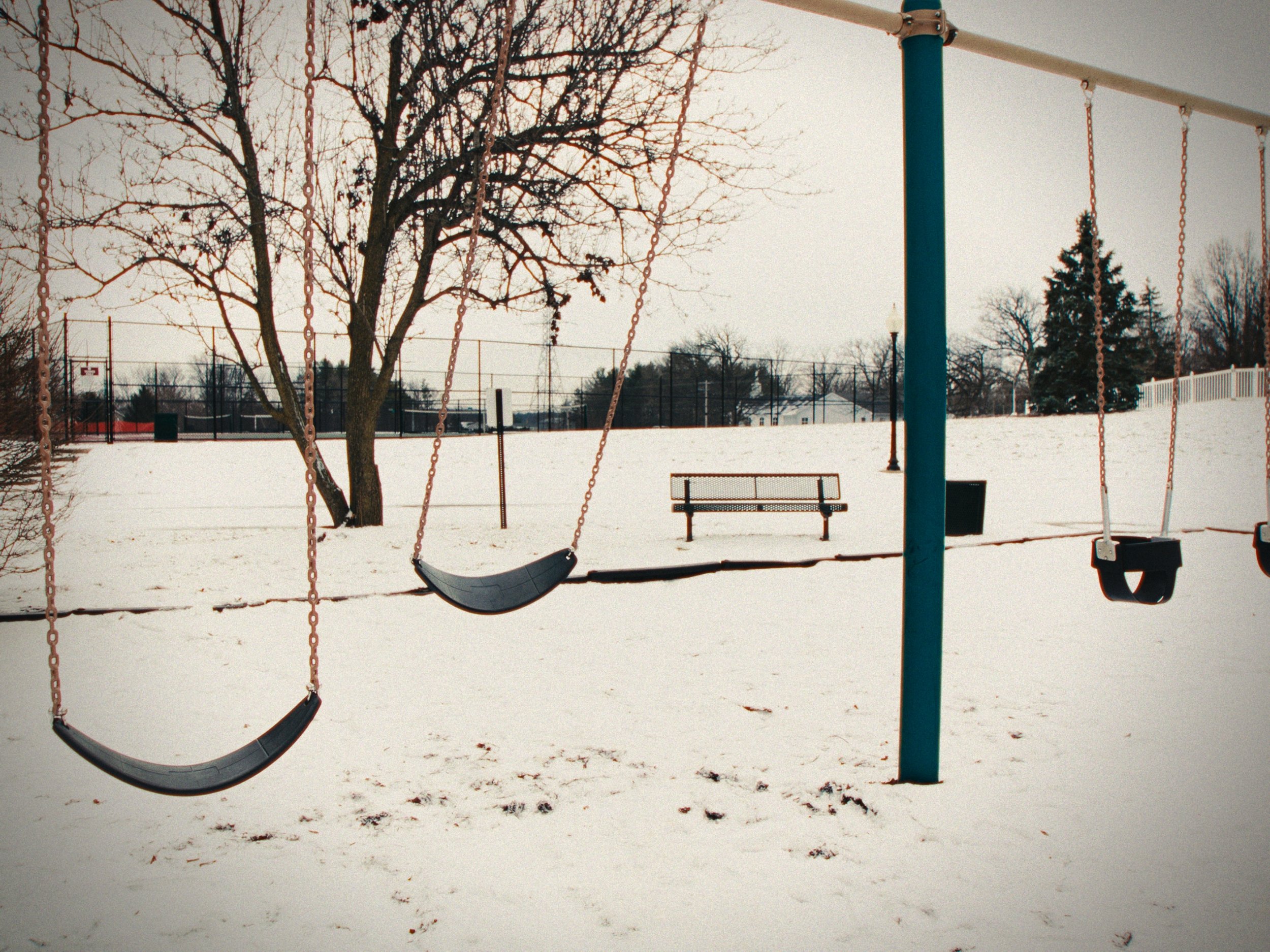 Empty swings on a snow-covered playground with a tree and a bench in the background.