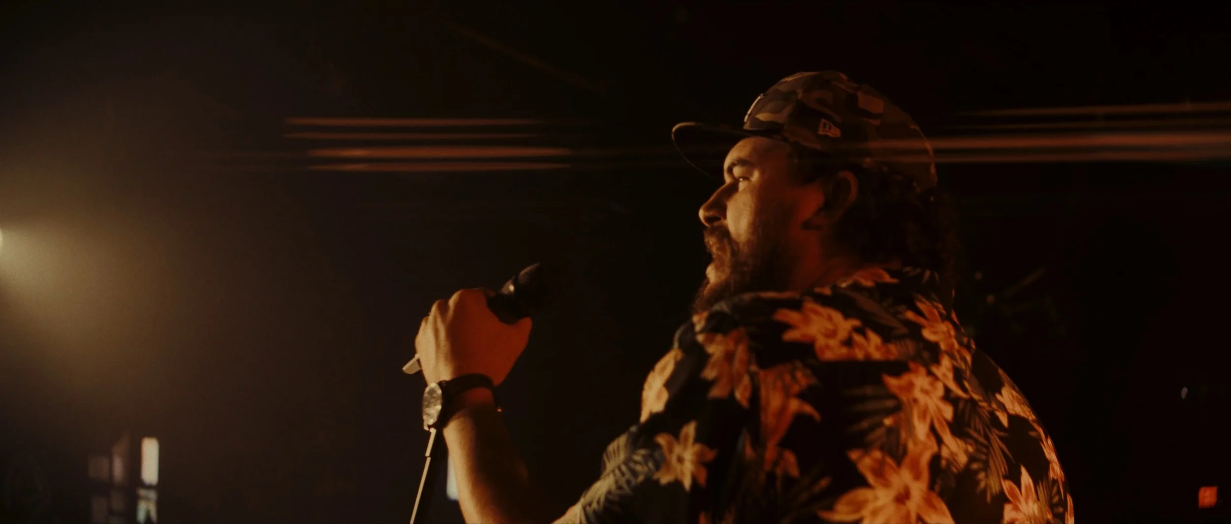 A man with long hair and a beard wearing a floral shirt and camouflage hat, holding a microphone and singing in a dimly lit environment.