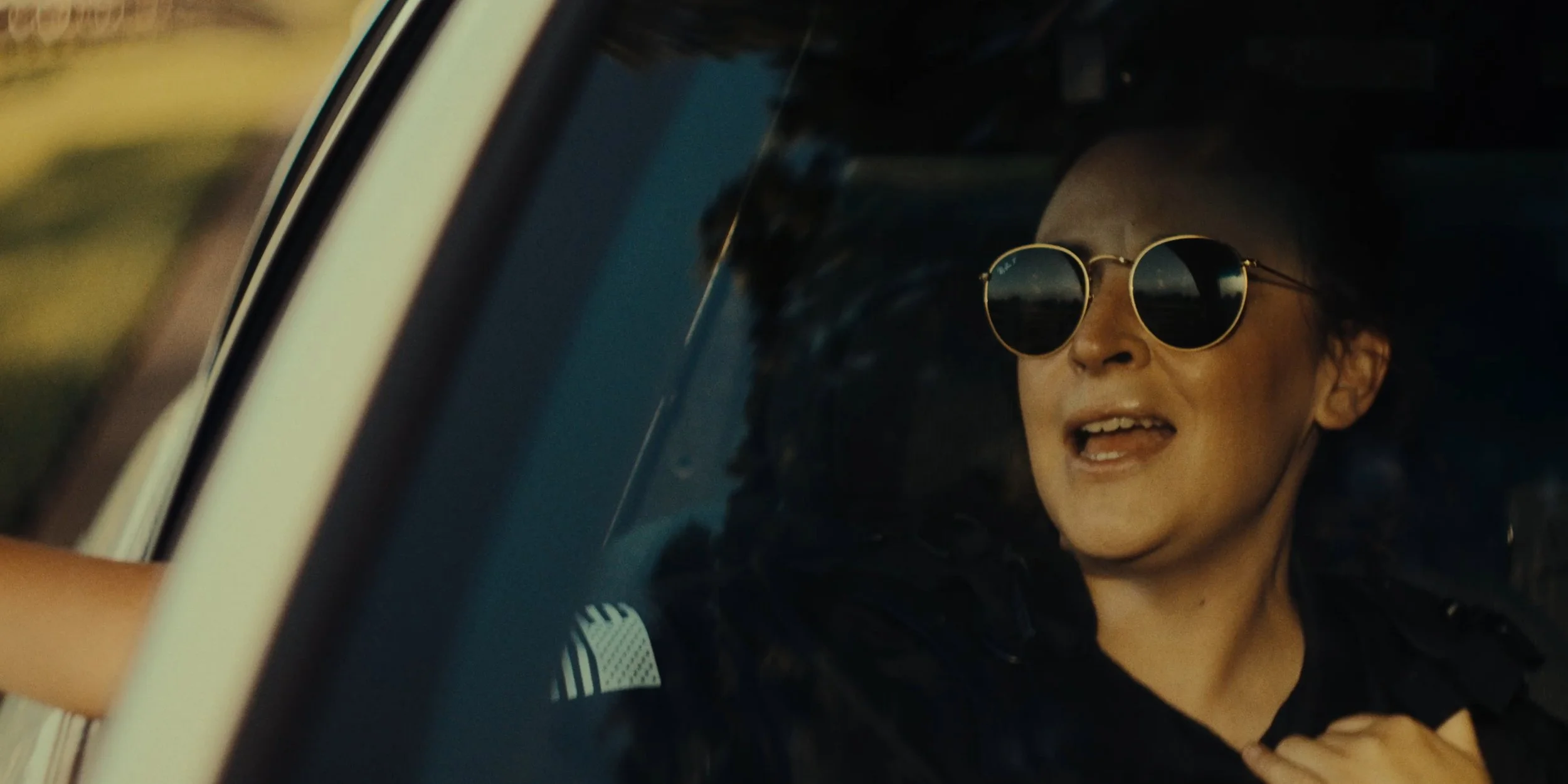 A woman wearing round sunglasses and a black jacket, driving a car with a joyful expression.