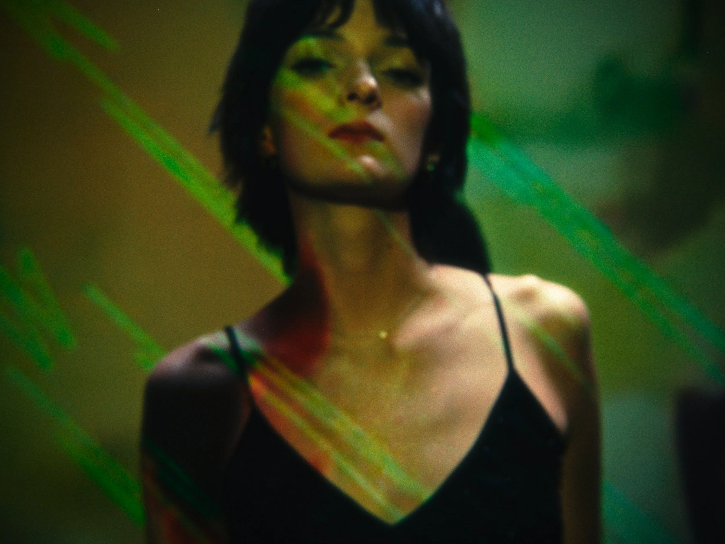 A woman with short black hair and dark lipstick, wearing a black spaghetti strap dress, standing in front of a green background with diagonal green light streaks.