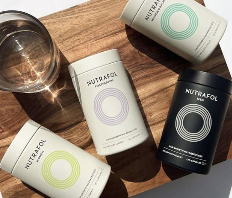 Four Nutrafol supplements in white, green, purple, and black bottles on a wooden surface with a glass of water