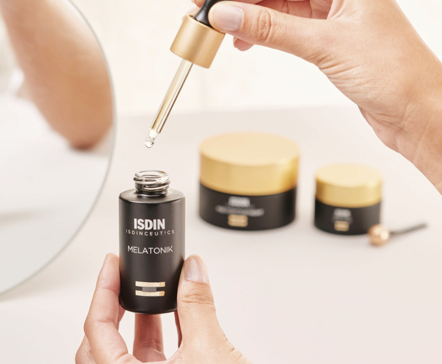 A person holding a black bottle labeled 'ISDIN Melatonik' with a dropper, with other skincare or cosmetic jars in the background on a white surface