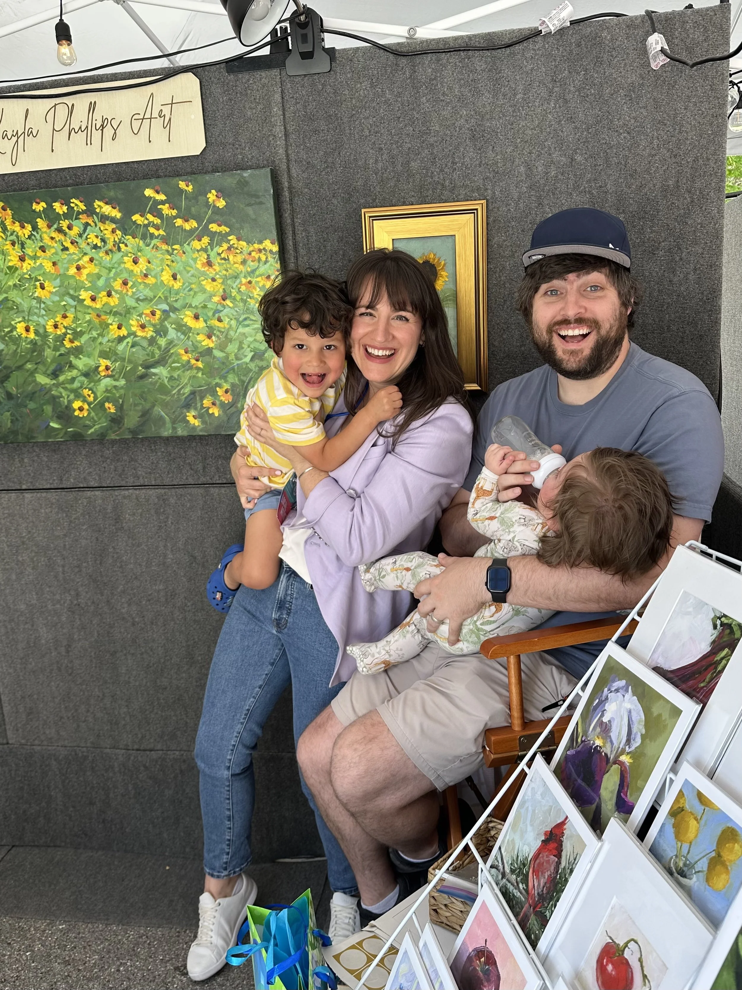 A family of four enjoying themselves at an art booth. The woman and  are smiling, with the  being held by the woman. The man and a young child sitting on his lap are also smiling at the camera. The background features paintings of flowers, including sunflowers, and framed artwork, with a display of small art prints of flowers and cherries in front.