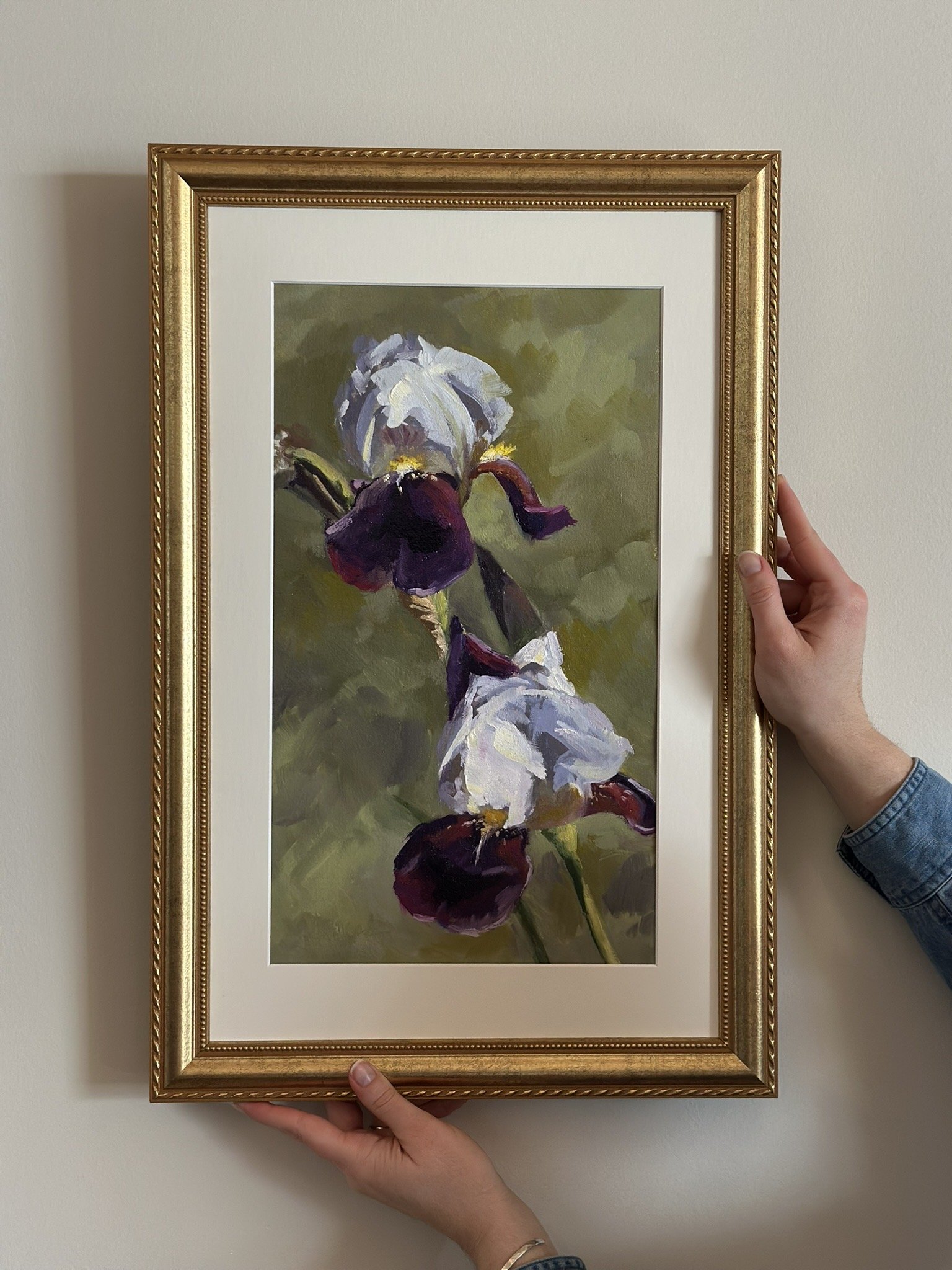 Painting of purple and white iris flowers in a gold frame, being held by two hands in front of a plain wall.