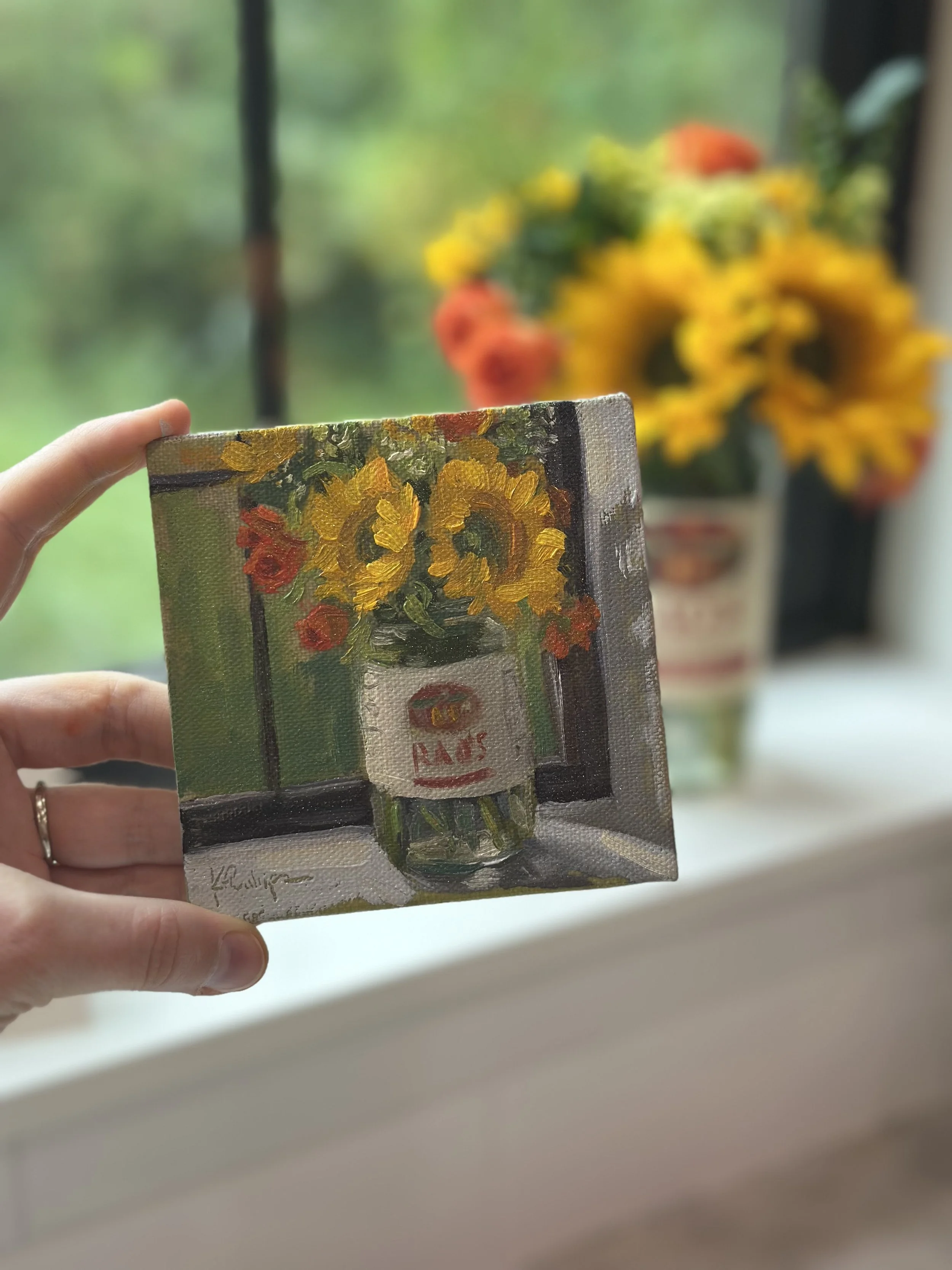 A small canvas painting of a bouquet of sunflowers in a jar, with a blurred background of a larger floral arrangement of sunflowers and orange flowers.
