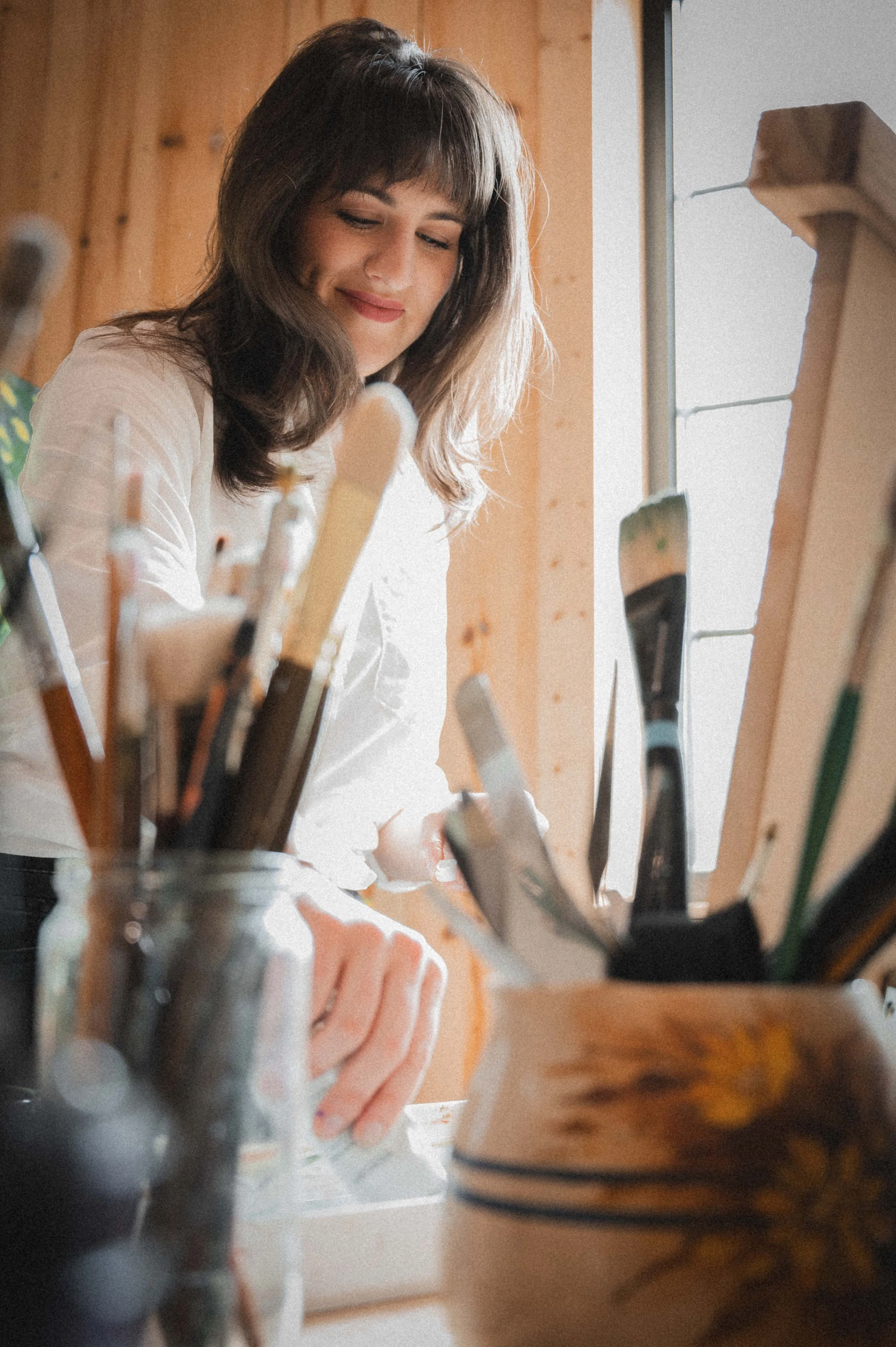 A woman with dark hair and a white shirt smiling while looking at her easel in an art studio with wooden walls and natural light from a window.
