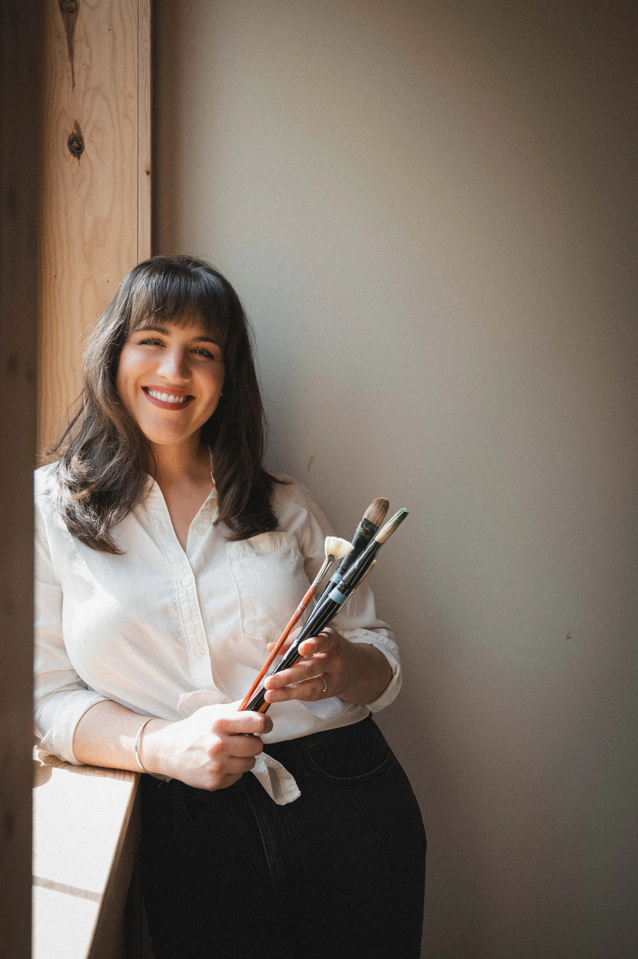Woman smiling and holding paintbrushes in a bright room with a wooden wall.