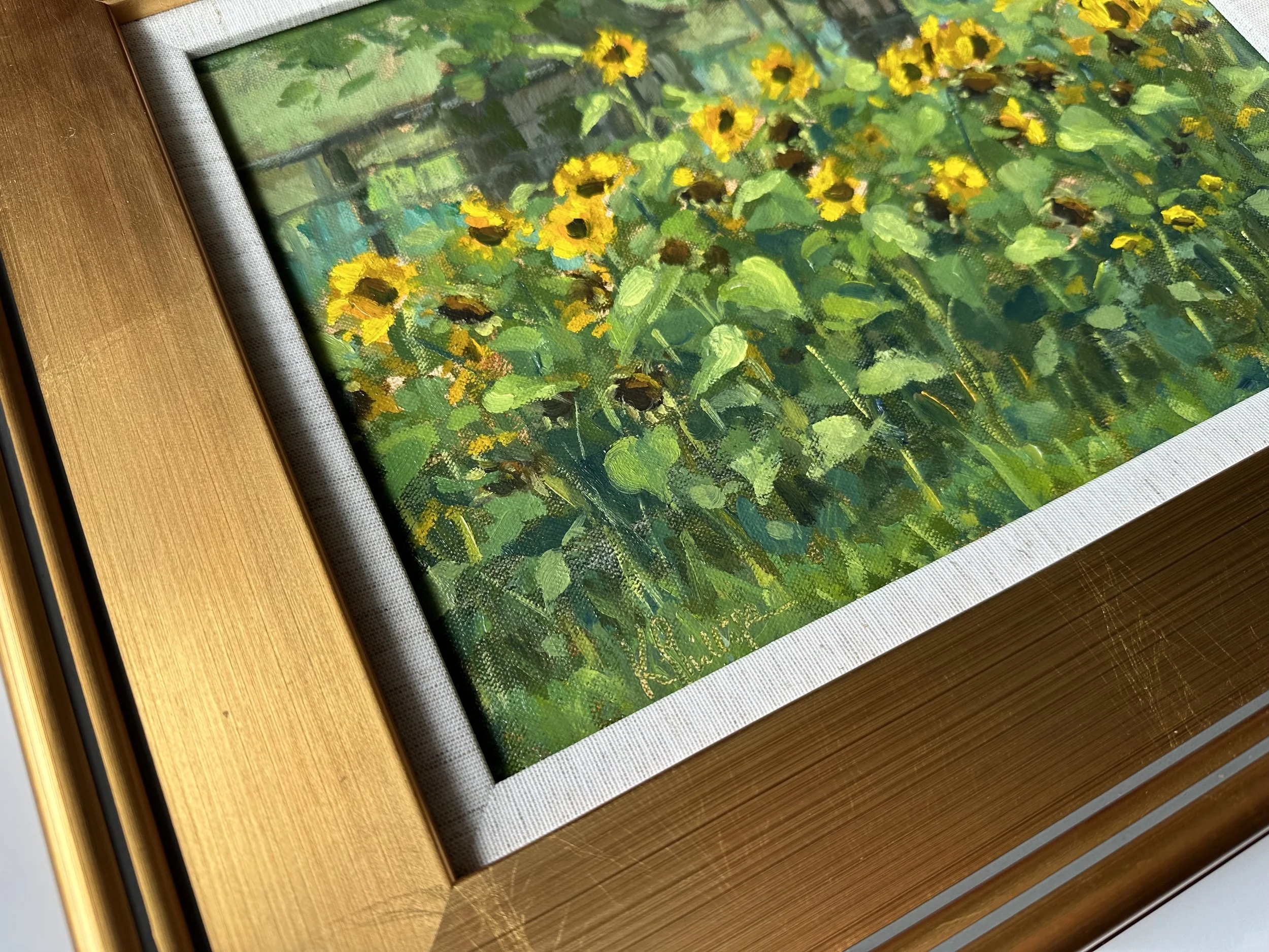 Close-up of a framed painting of yellow sunflowers in a garden, with a signature at the bottom.