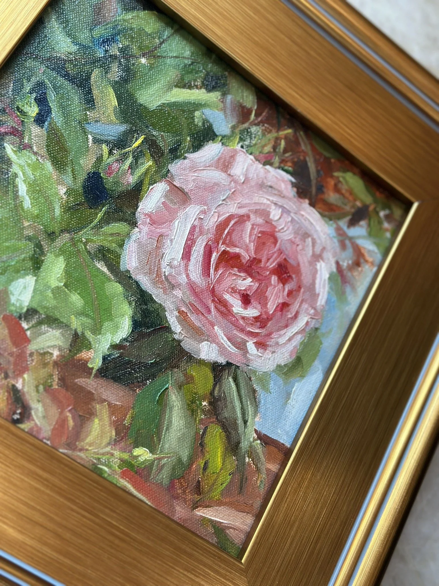 Close-up of an oil painting of a pink rose surrounded by green leaves, framed in a wooden frame with a gold inner edge.