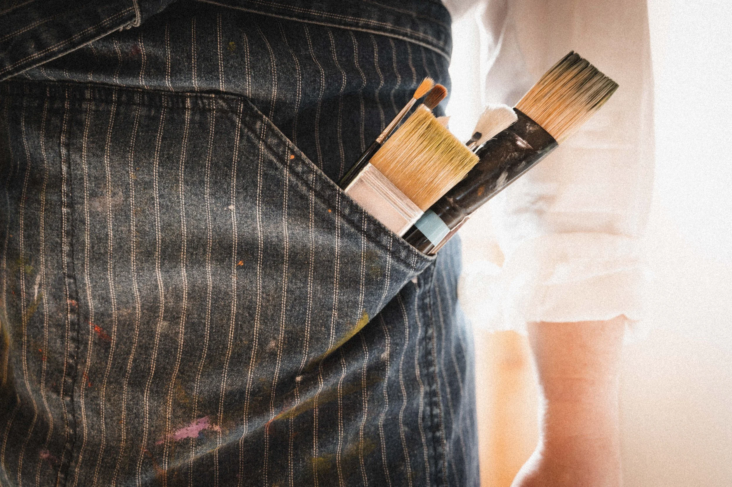 Close-up of a carpenter's pocket with various paintbrushes and tools inside, worn denim work pants with paint stains.