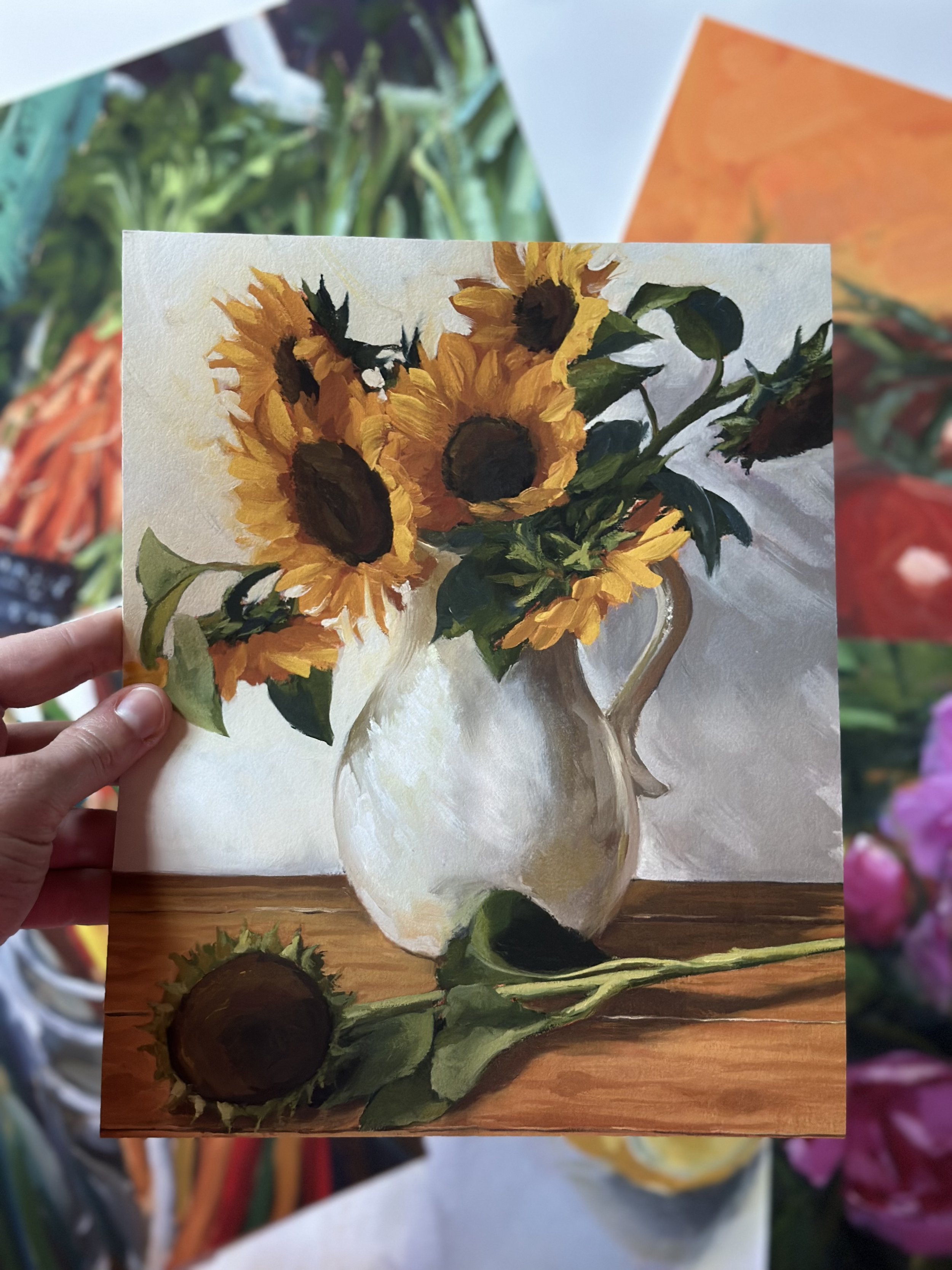 A painting of a bouquet of sunflowers in a white ceramic pitcher on a wooden table, with a sunflower lying on the table in front of the pitcher.
