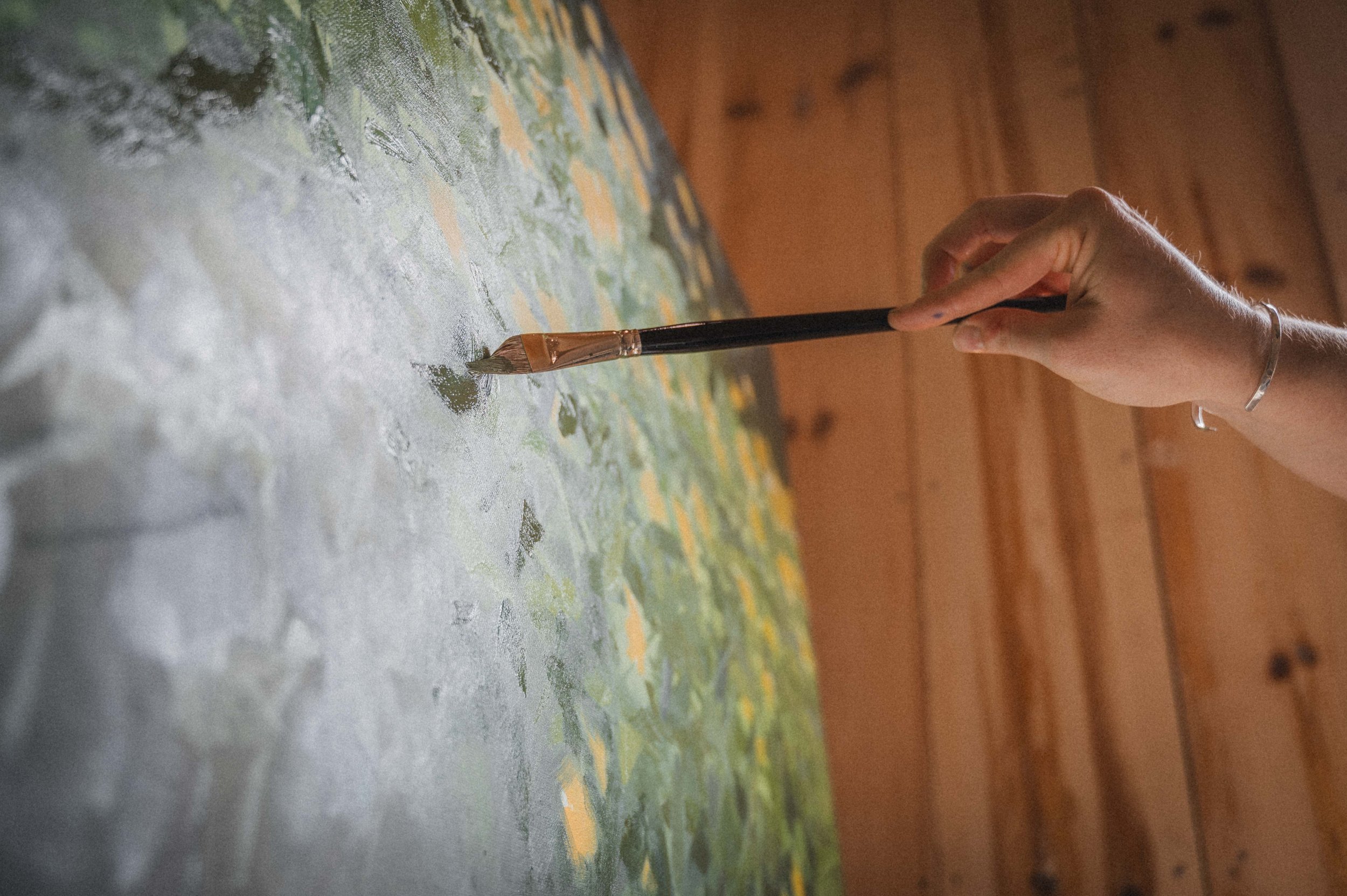 Close-up of a person's hand holding a paintbrush, painting on a textured canvas with abstract green, yellow, and white patches, with a wooden wall in the background.