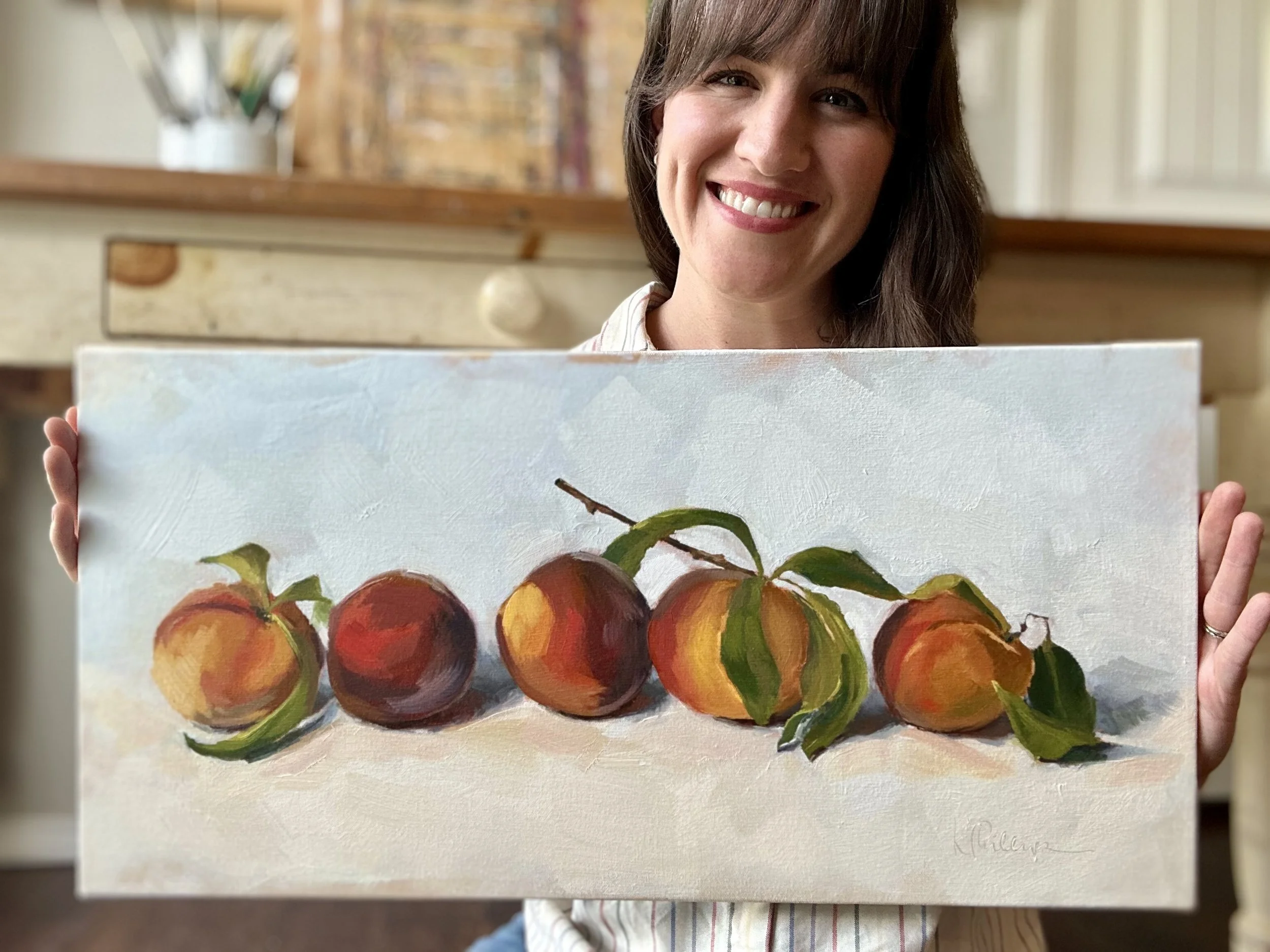 A woman smiling and holding a painting of five peaches with green leaves on a light background.